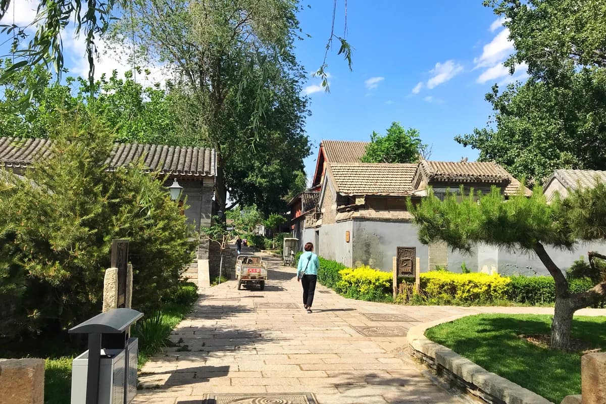 Sunny Beijing hutong lane with courtyard houses and a pedestrian walking through the neighborhood.