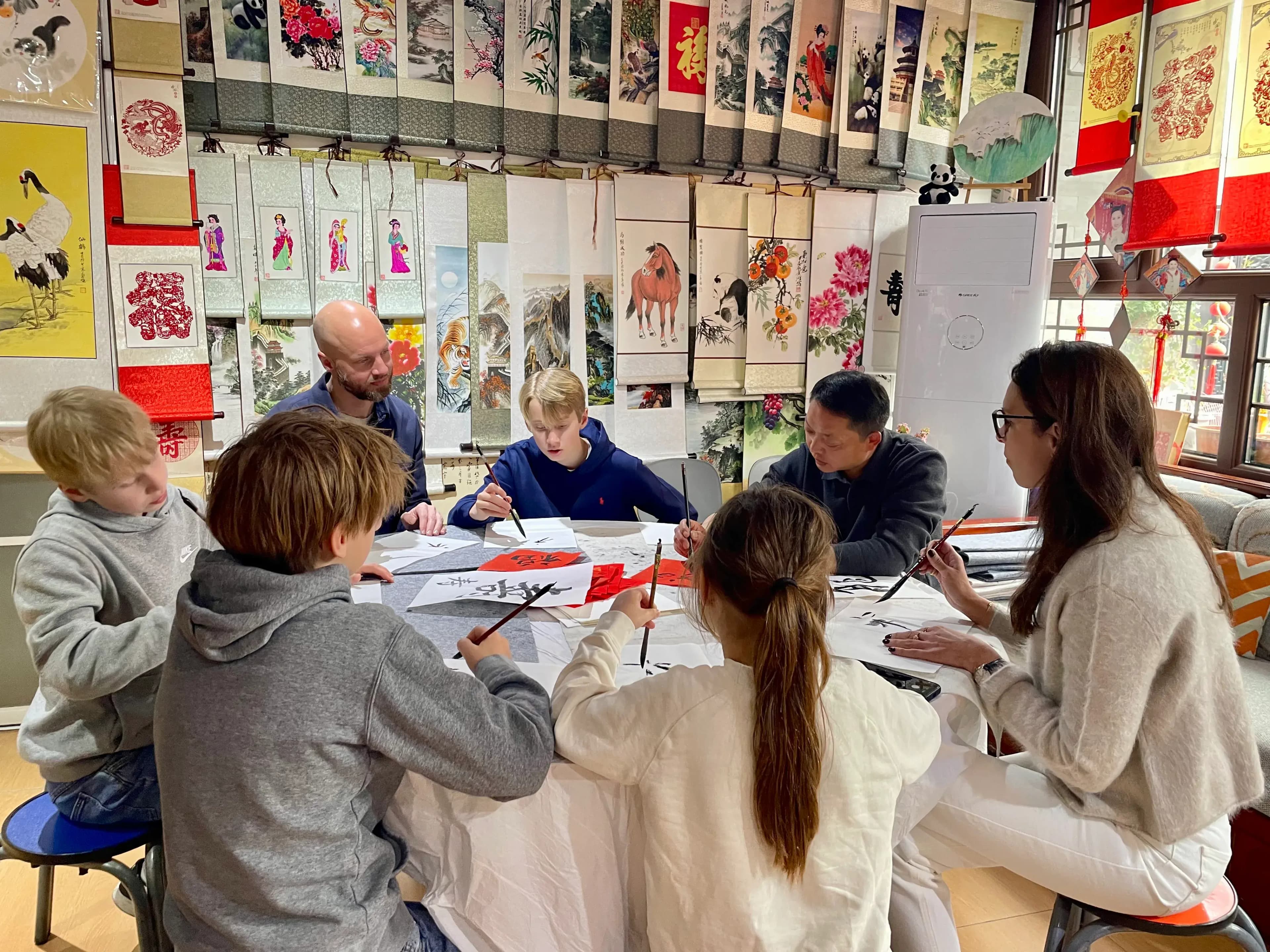 A group of people, including children and adults, are seated around a table practicing calligraphy with brushes and ink.