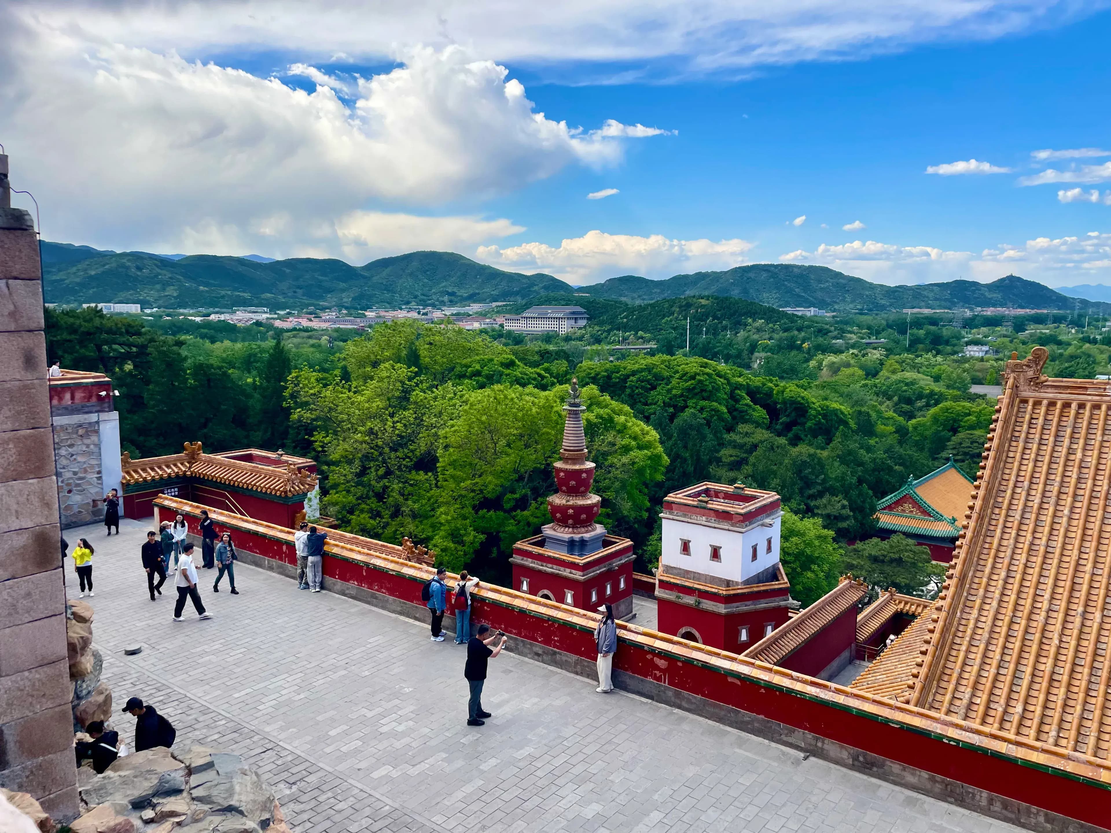 Aerial view of the Four Great Regions at the Summer Palace, showing traditional Chinese architecture with red and gold details, surrounded by lush green trees and hills in the background.