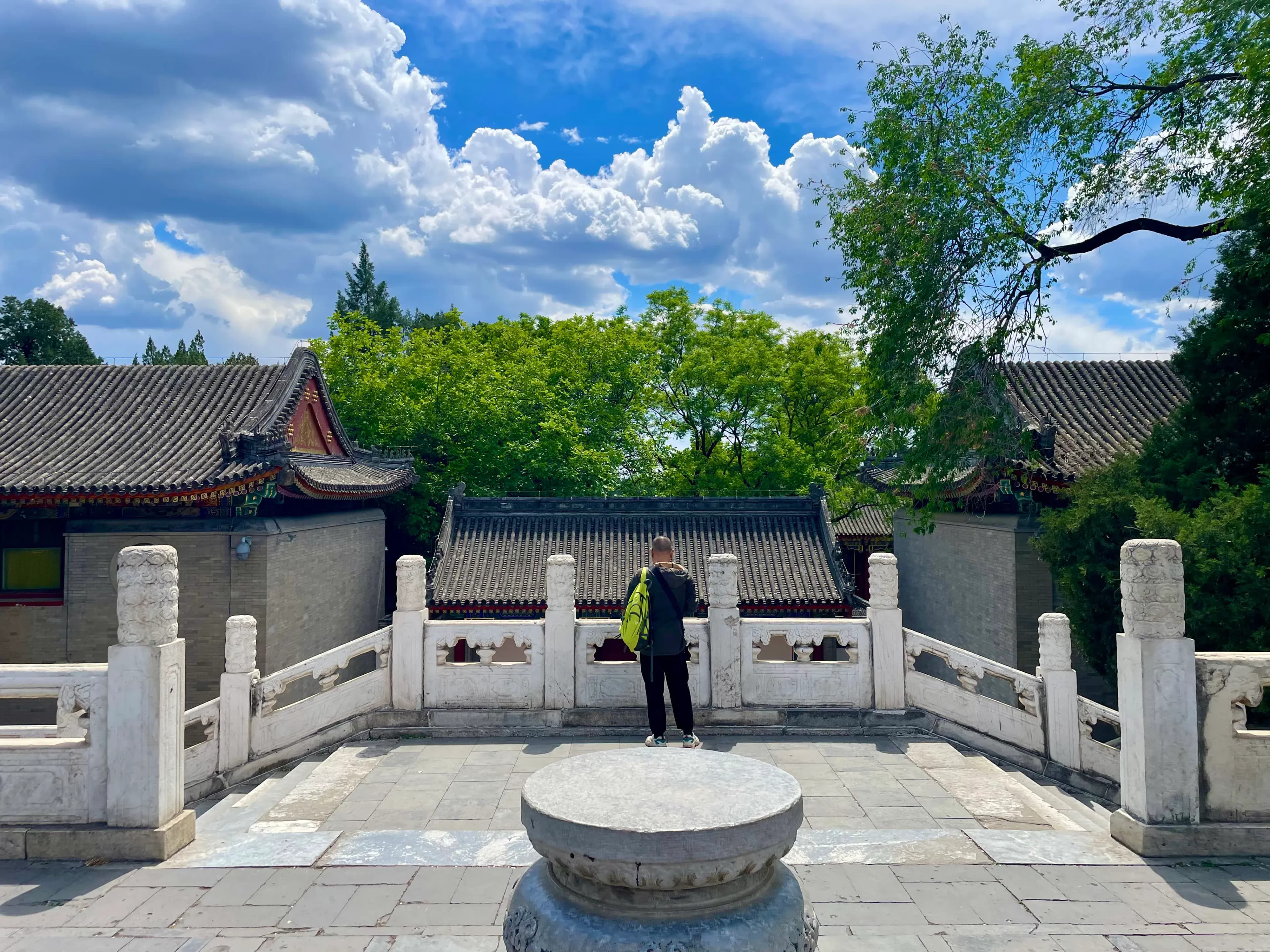A person stands on a stone platform overlooking traditional Chinese buildings with tiled roofs and lush green trees under a bright blue sky with clouds.