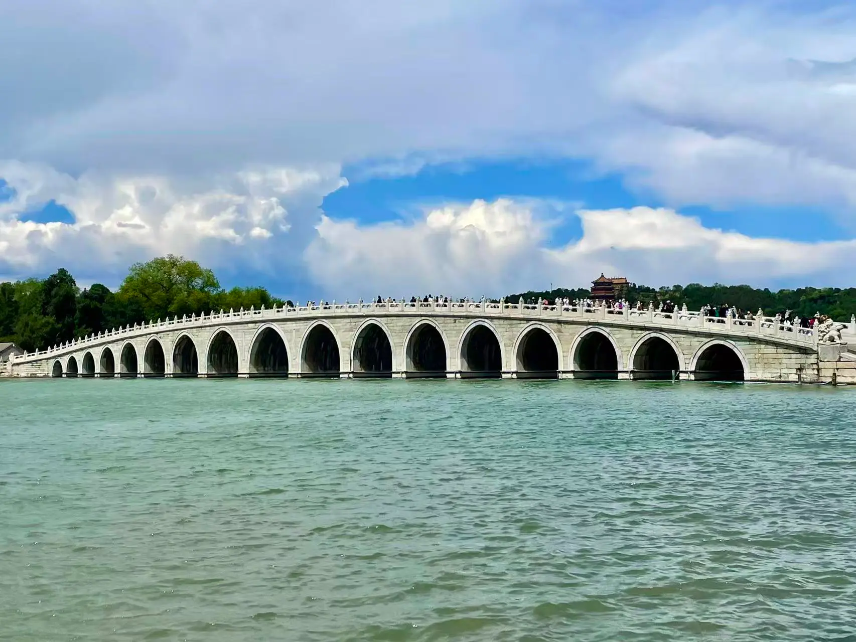 A long stone bridge with seventeen arches spans across a body of water, with people walking on it.