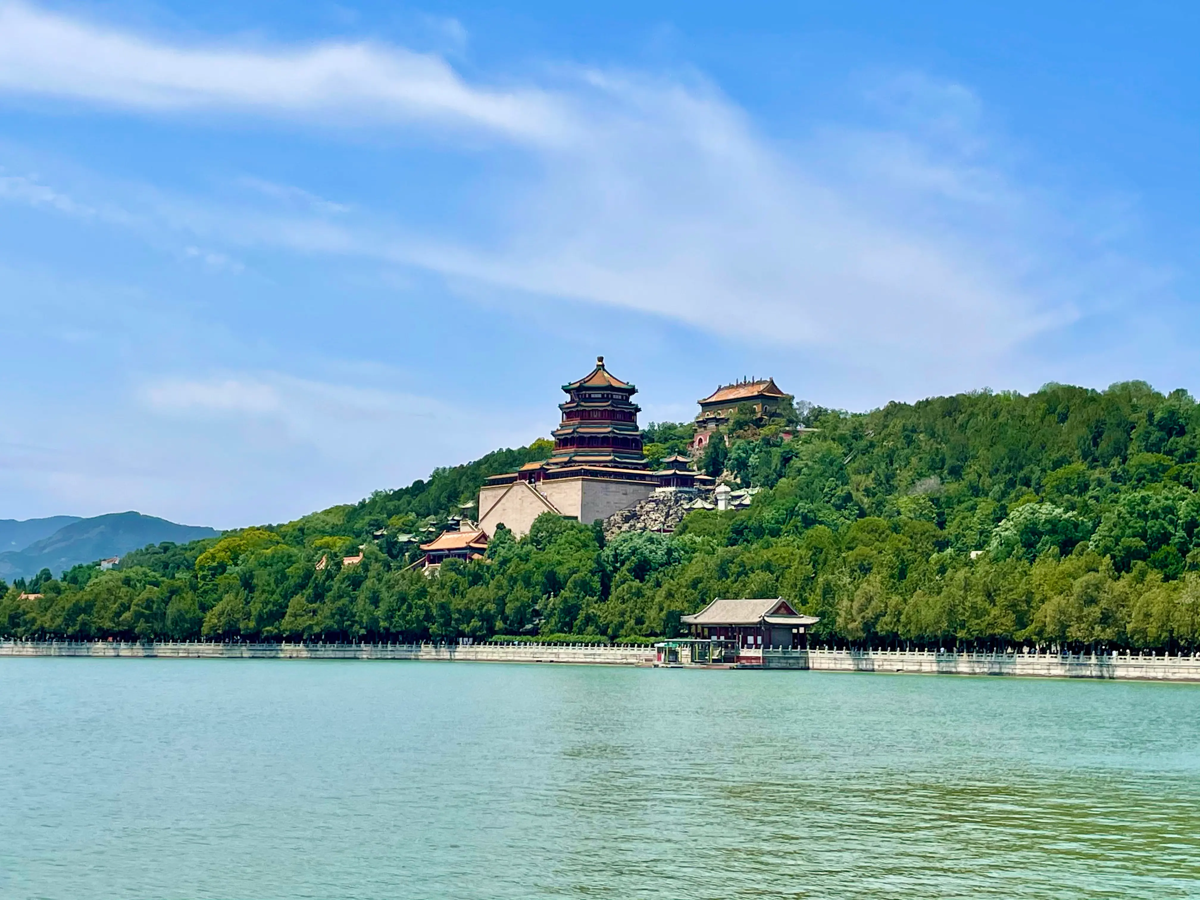 The Tower of Buddhist Incense is situated atop a lush green hill, overlooking a serene body of water under a clear blue sky.