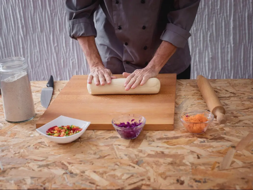 A chef kneading dumpling dough on a wooden cutting board with vegetables nearby