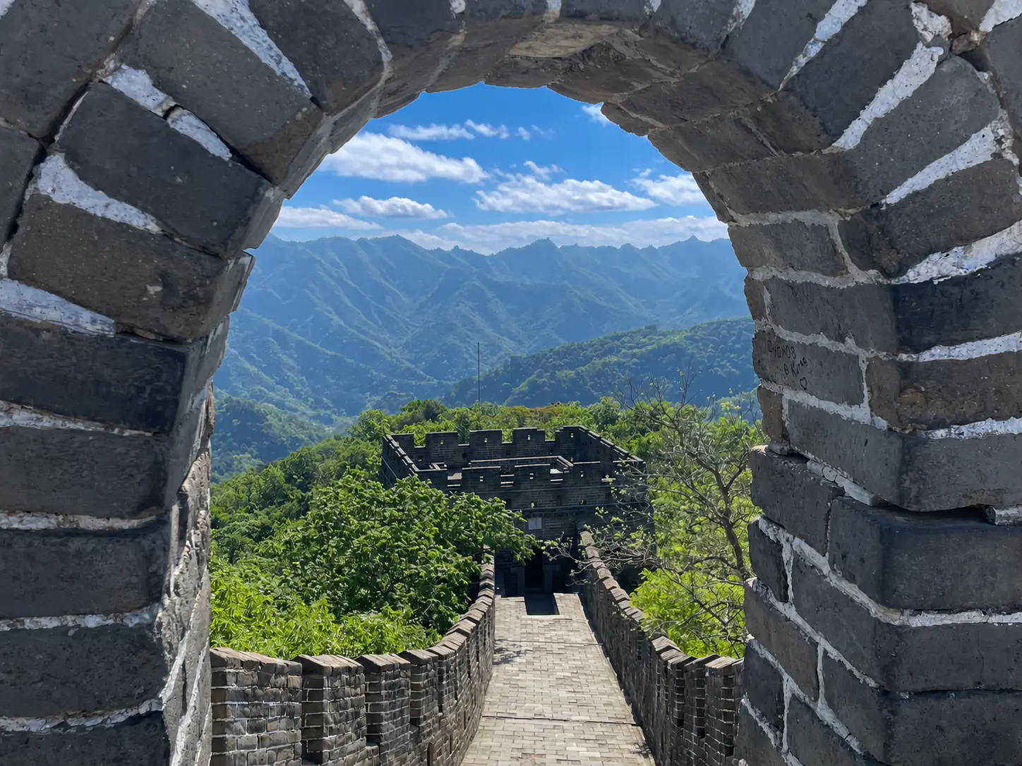 A stone archway frames a view of the Great Wall stretching into the distance, with lush green mountains and a clear blue sky in the background.