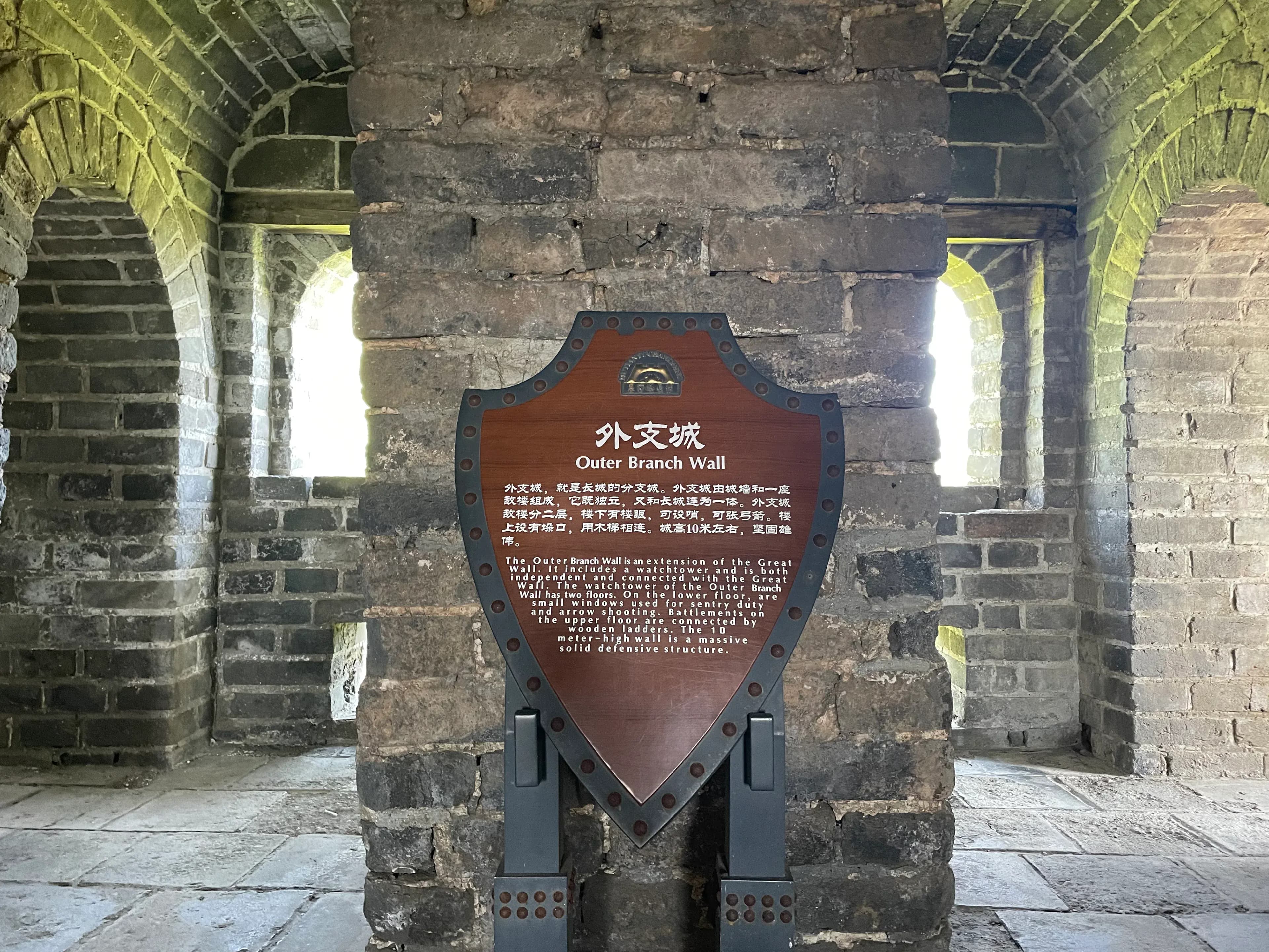 A wooden sign with information about the Outer Branch Wall, set against a stone wall with arched windows.