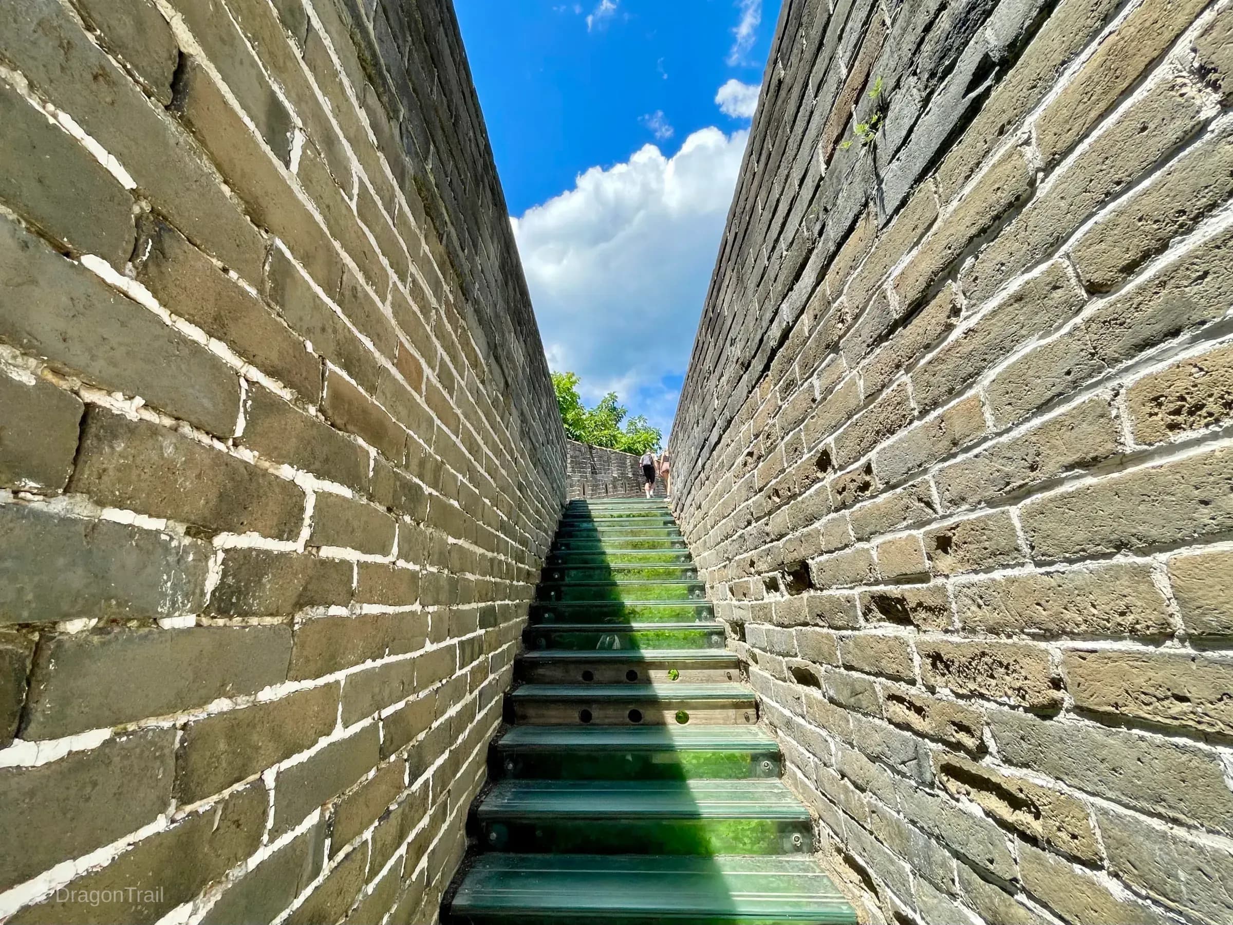 Low-angle shot looking straight up green translucent glass steps between tall Mutianyu brick walls toward hikers and blue sky