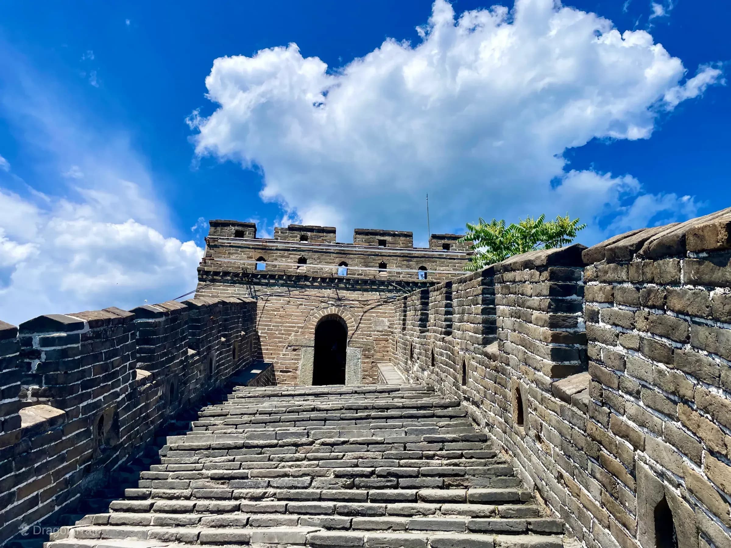 Low-angle view up broad Mutianyu stone stairs into Tower 10’s single central arched doorway against a bright cloudy sky