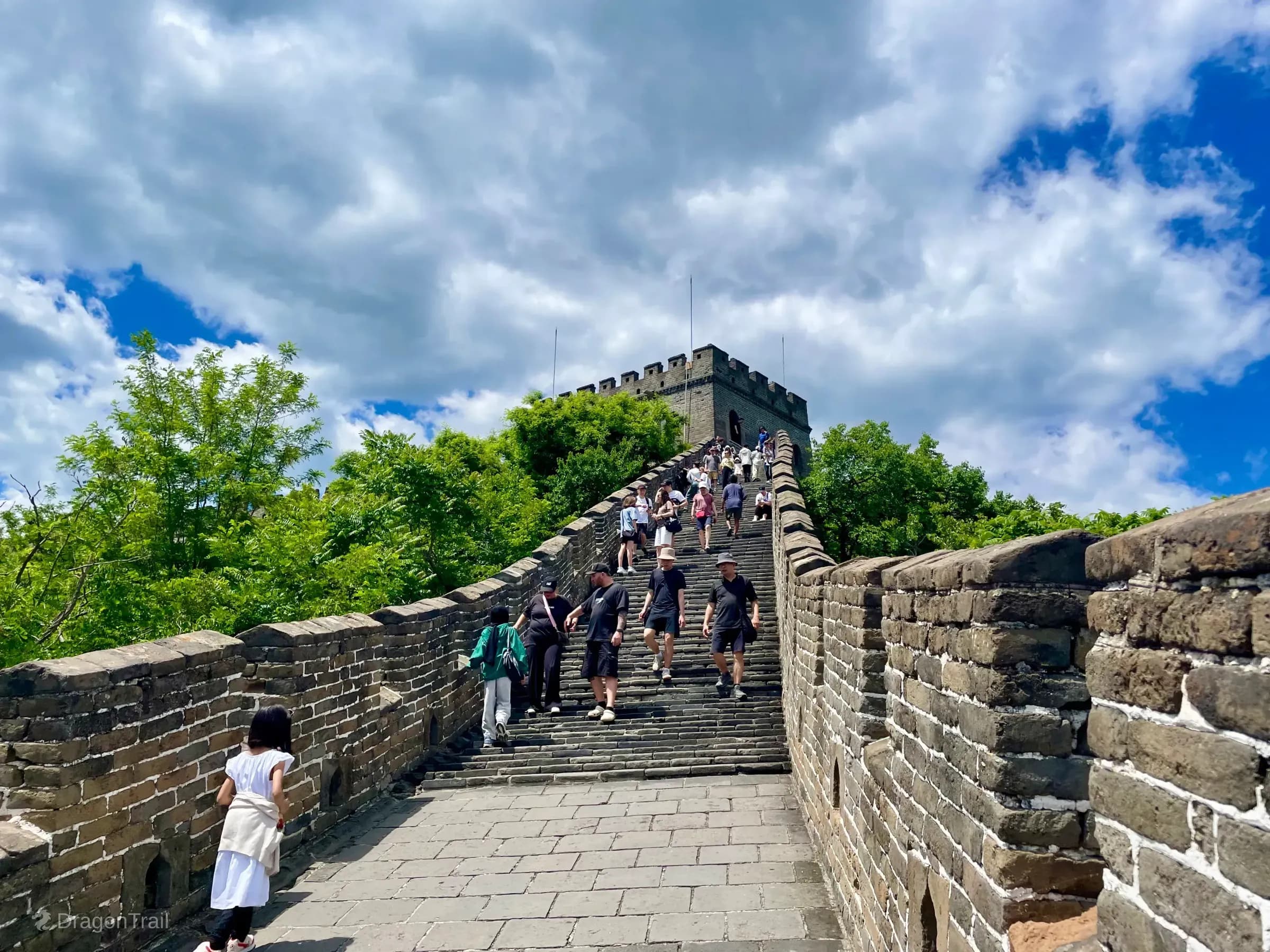High-angle view up a steep Mutianyu stairway packed with tourists climbing toward Tower 14’s battlemented watchtower under a blue sky with white clouds