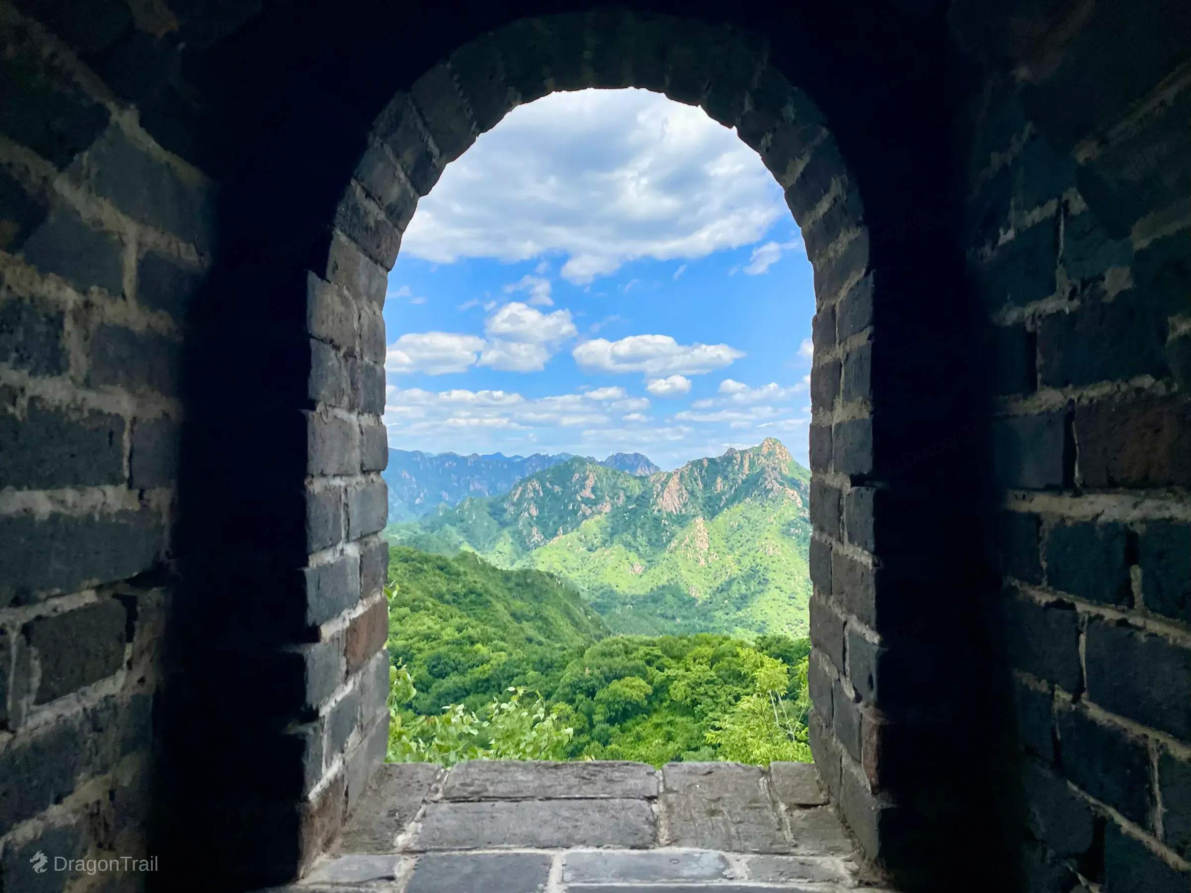 View from inside Mutianyu Tower 15 through a dark semicircular brick arch framing lush green mountains and a bright blue sky with scattered clouds