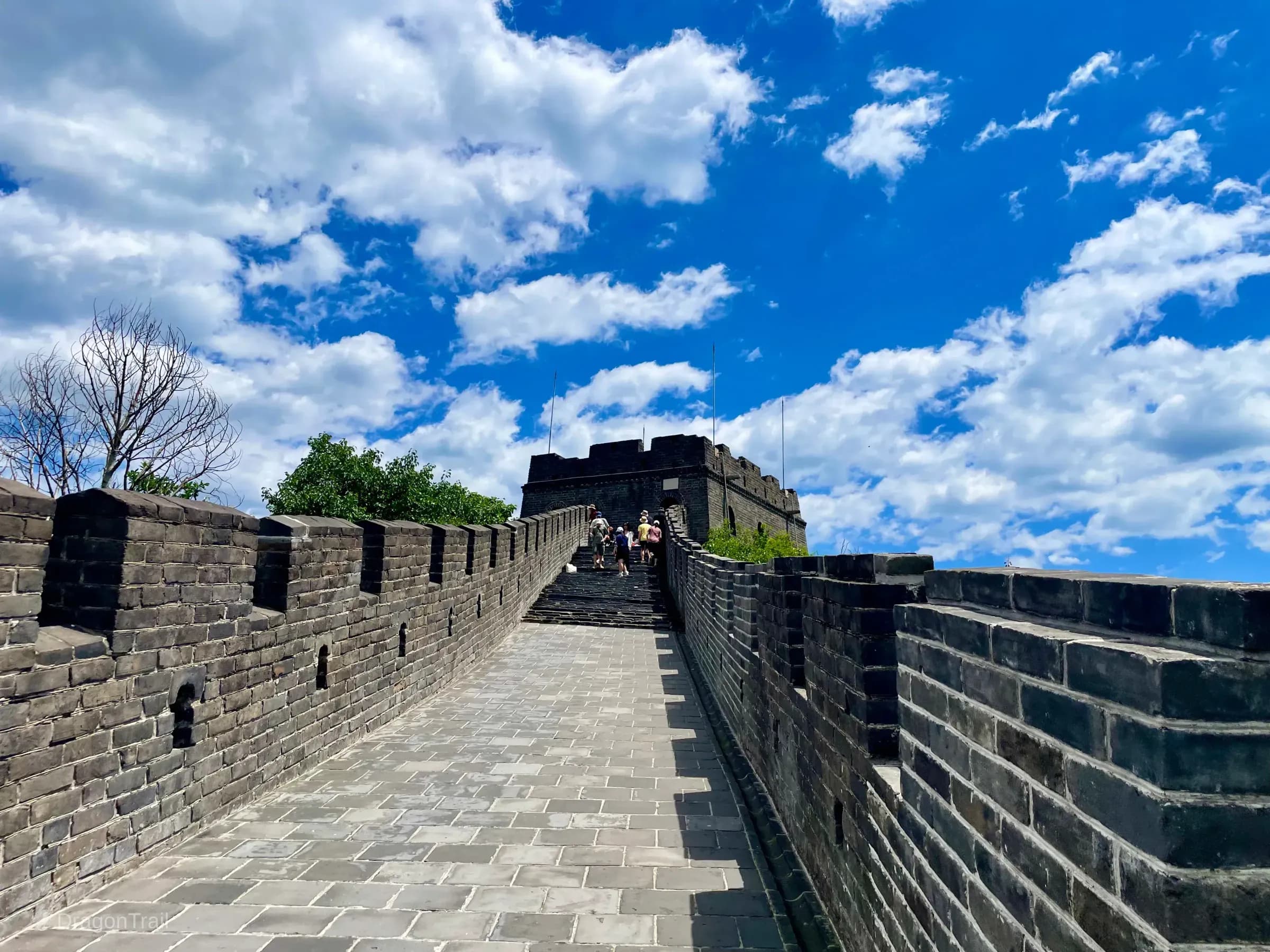 Looking up Mutianyu’s stone walkway and stairs toward Tower 15’s battlemented watchtower, with tourists ahead, crenellated parapets, and a blue sky with white clouds