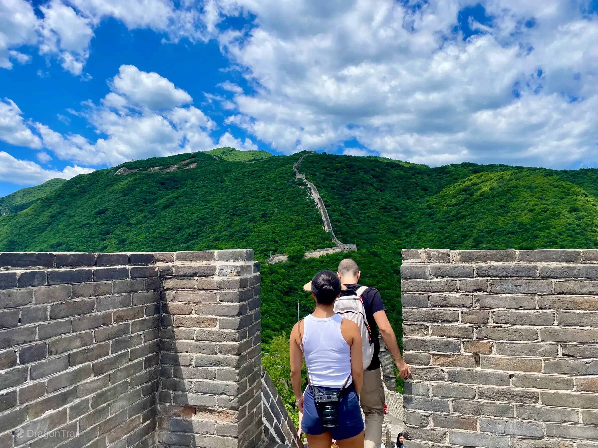 Two visitors look through a Mutianyu battlement gap toward a steep forested ridge where the Great Wall climbs past successive watchtowers under a cloudy blue sky