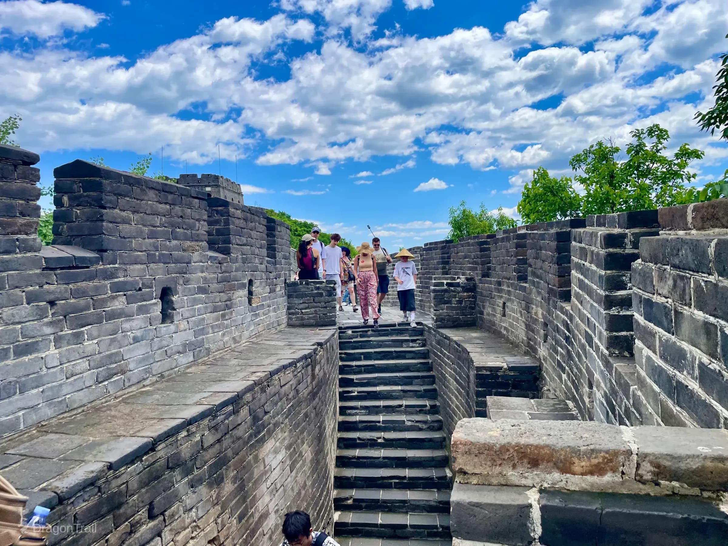 Looking down Mutianyu’s crenellated stone walkway and central stairs as tourists climb between wall levels, with green foliage over the parapets and blue sky above