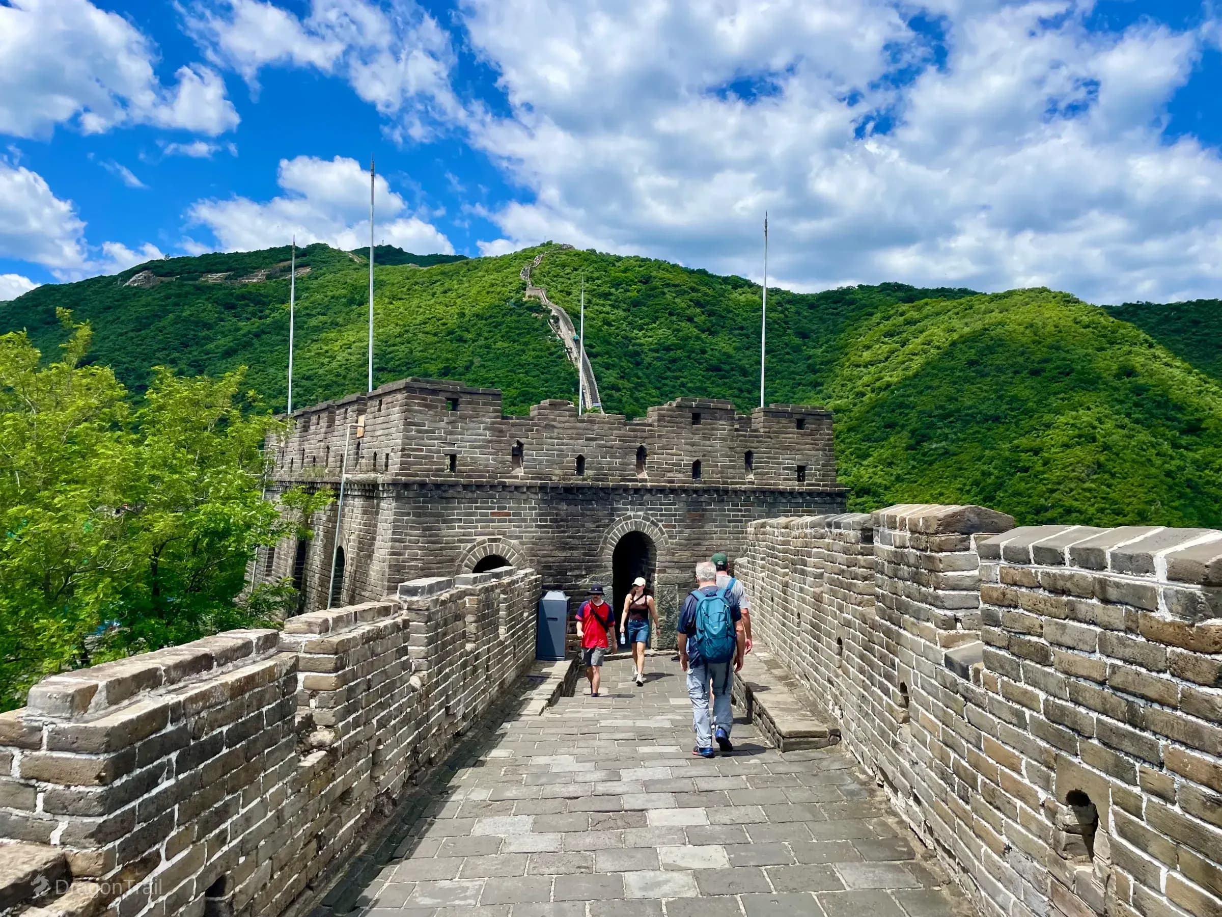 Wide Mutianyu stone walkway leading to Tower 17’s arched watchtower passage, with tourists ahead and the steep upper ridge wall climbing into green mountains beyond