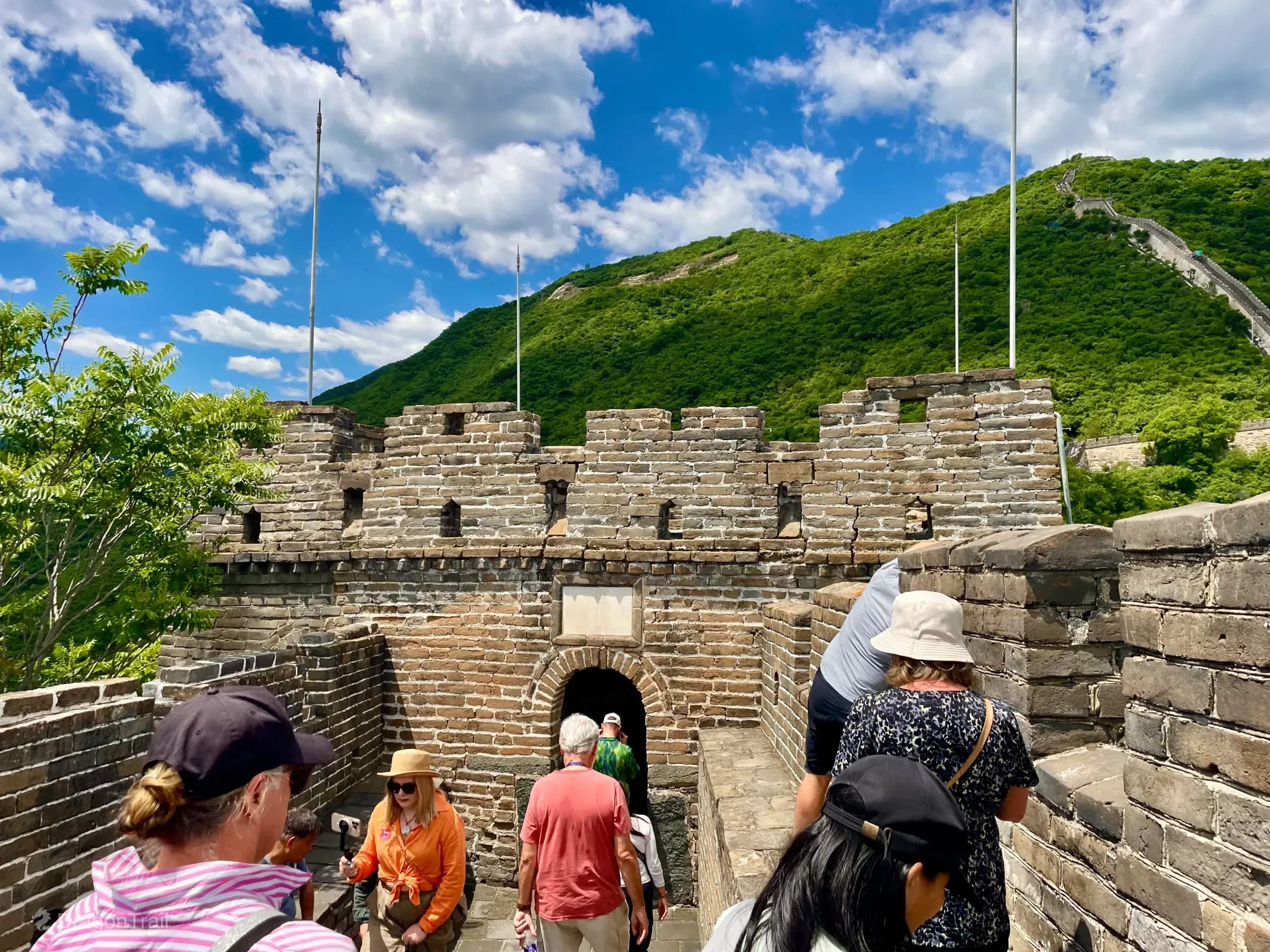 Tourists walking through Mutianyu Tower 18’s arched stone passage beneath a white plaque, with a large green mountain and distant ridge wall under a blue sky