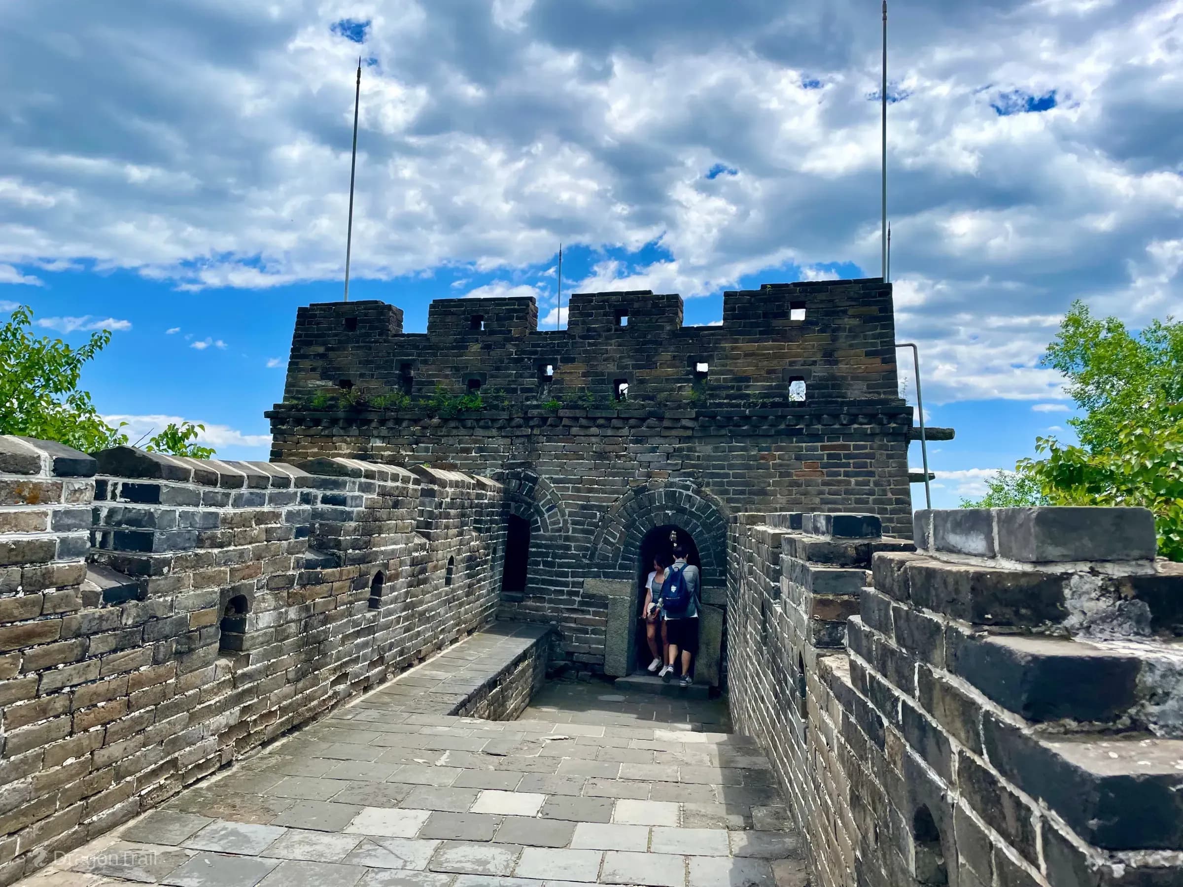 Looking along Mutianyu’s stone walkway to Tower 18’s square grey-brick watchtower with an arched doorway, crenellated parapet, lightning poles, and green foliage framing the scene