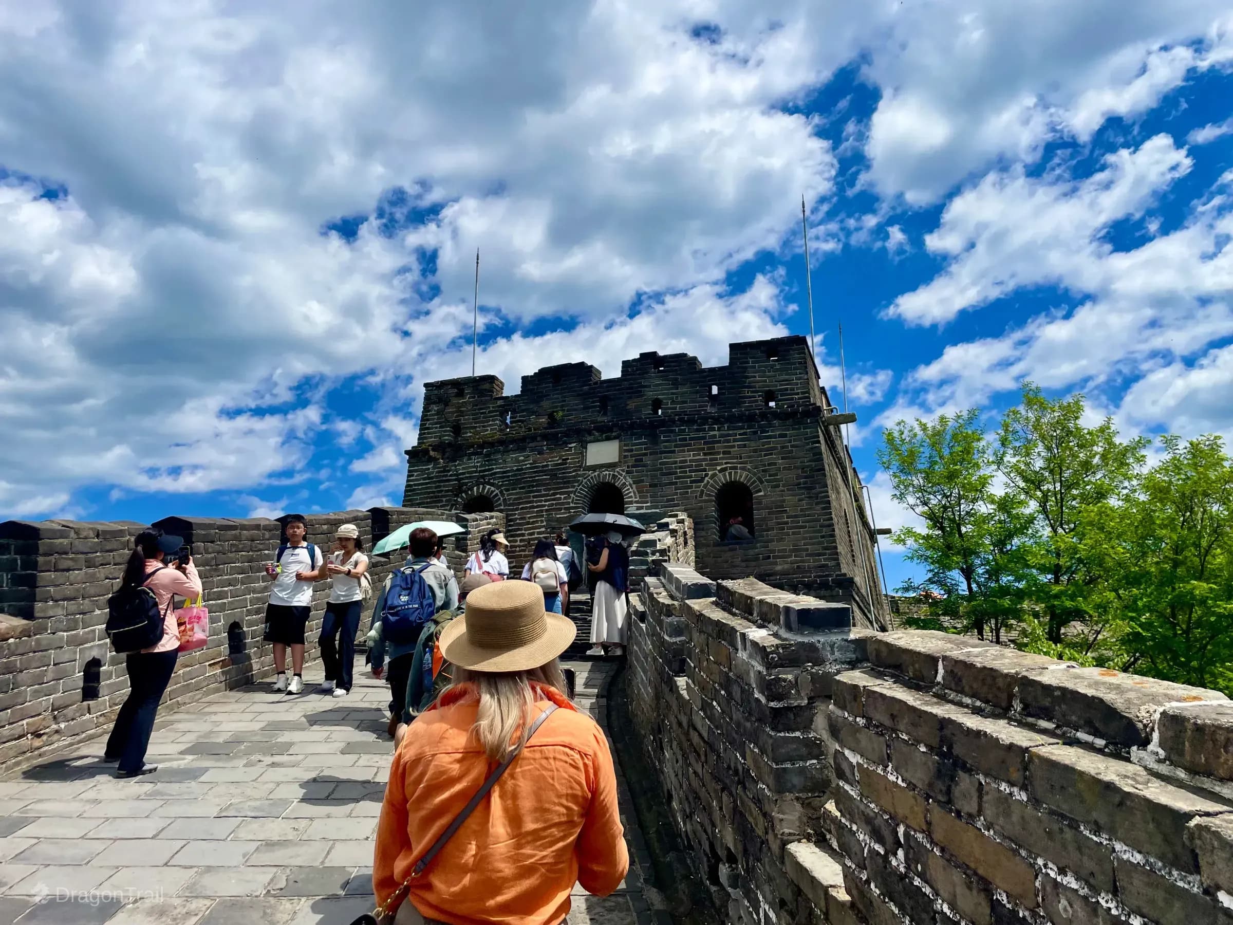 Tourists on Mutianyu’s stone walkway approaching Tower 18’s dark grey watchtower with arched openings, a white plaque above the central arch, and a cloudy blue sky
