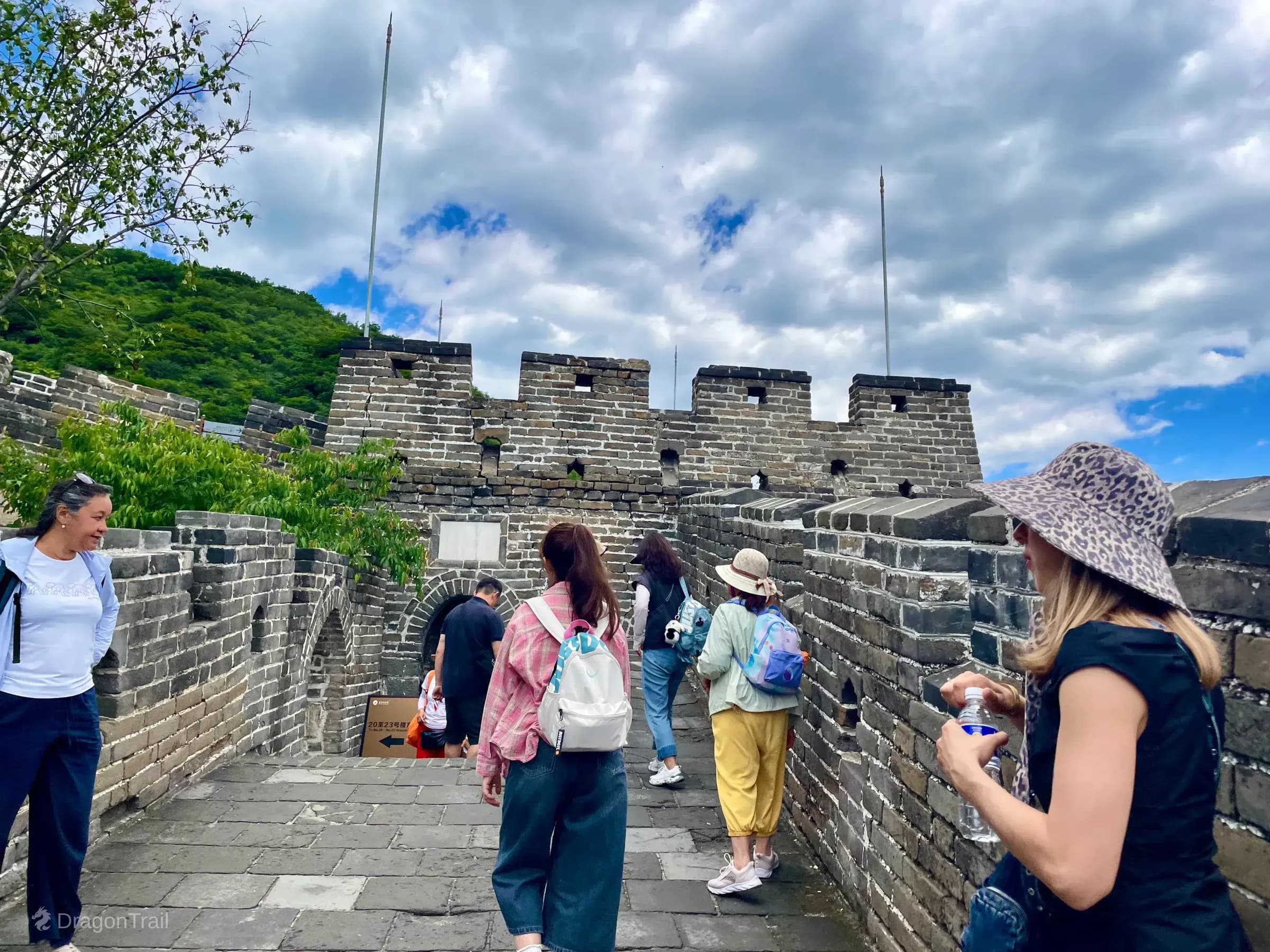 Tourists on Mutianyu’s stone walkway near Tower 19’s fortified crenellated section and arched openings, with forested hillside and cloudy sky