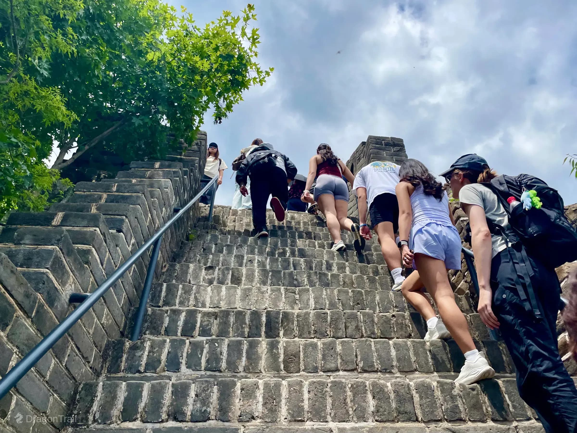 Low-angle view up Mutianyu’s steep Hero Slope stone stairs with a metal handrail as tourists climb toward Tower 20’s battlements under a bright cloudy sky
