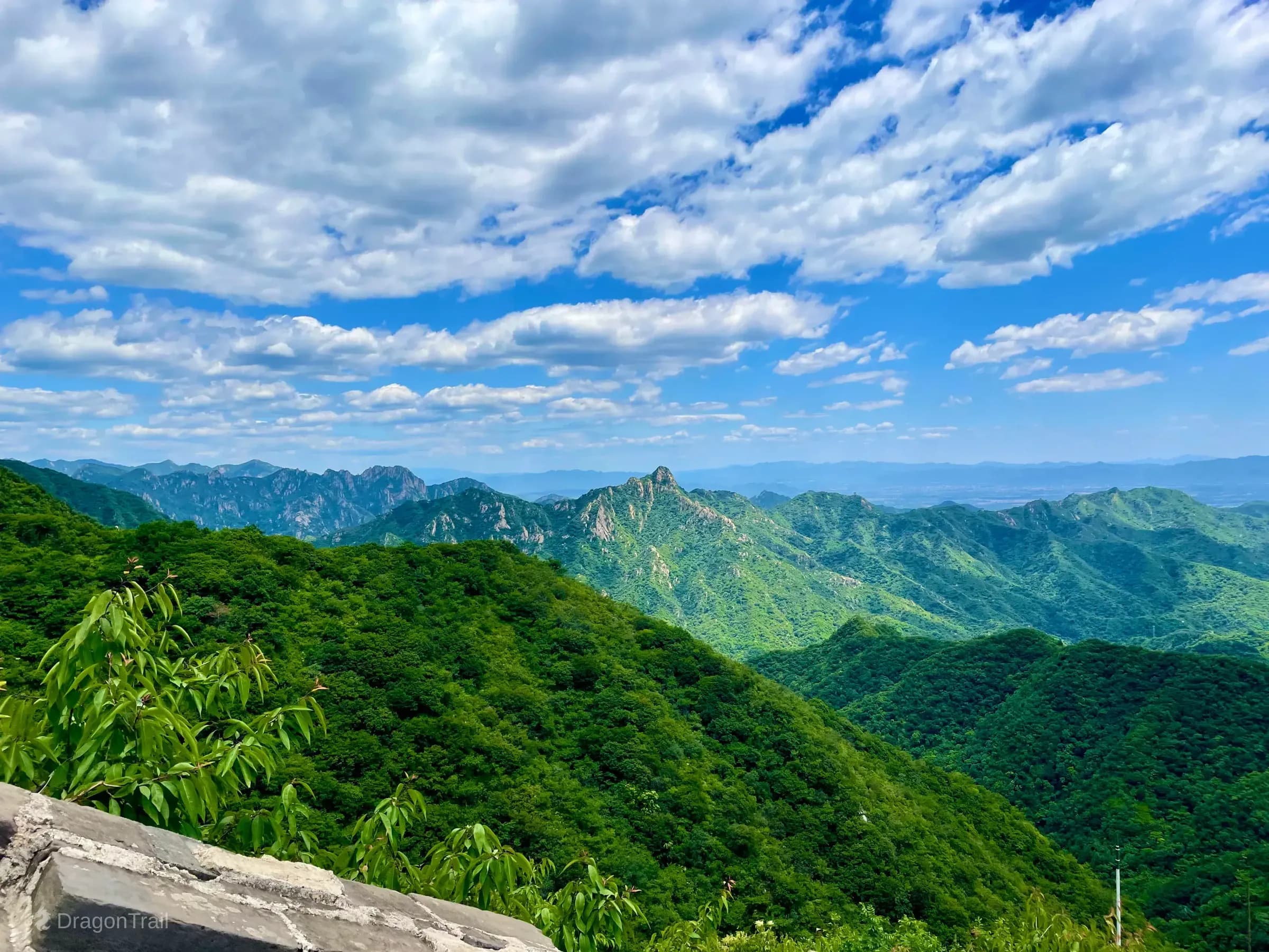 Panoramic view from Mutianyu Tower 21 over densely forested mountain ridges fading to blue haze, with a grey stone parapet corner and green leaves in the foreground under a bright cloudy sky