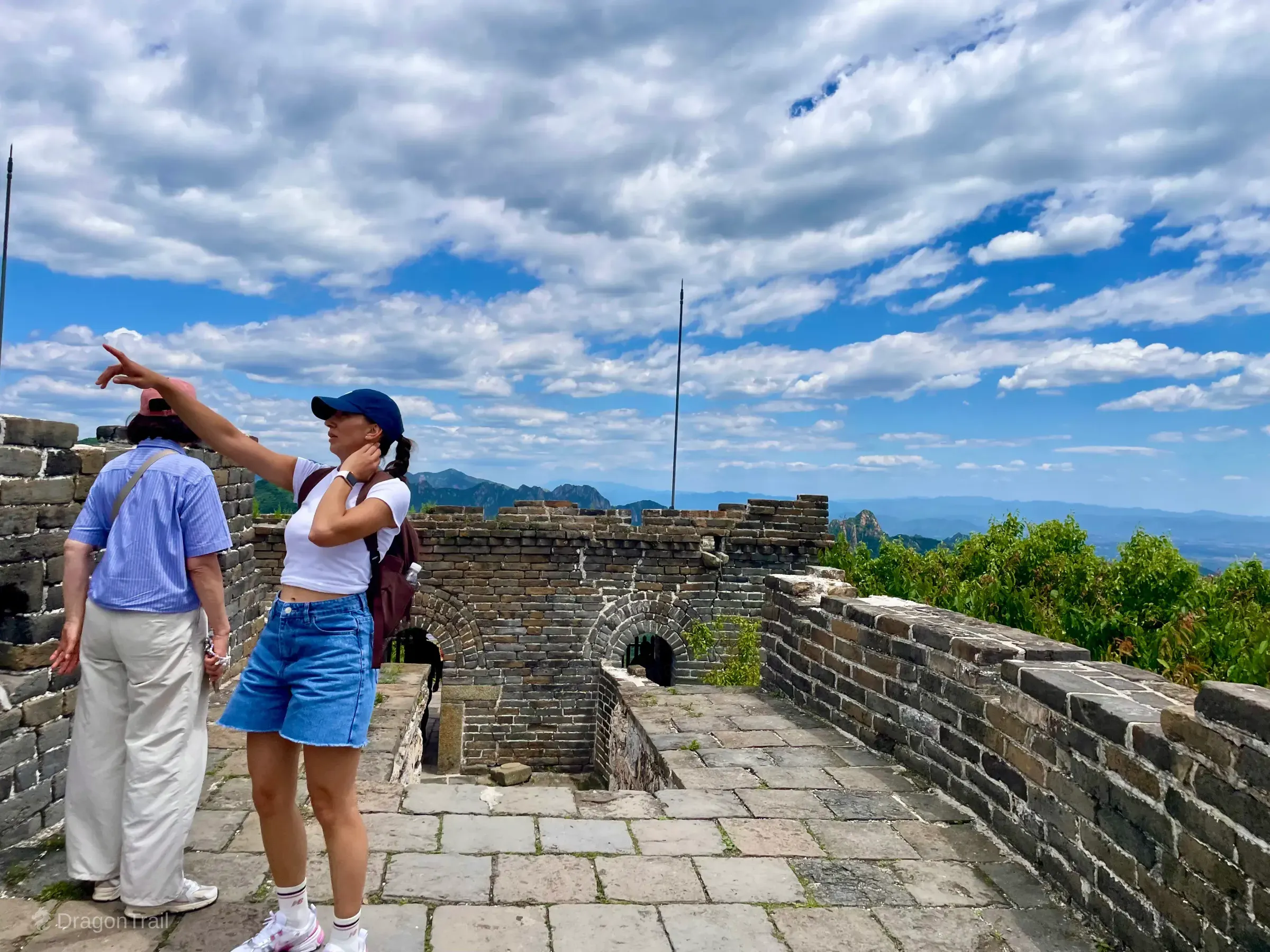 Two tourists on Mutianyu Tower 21’s stone rooftop platform pointing toward rolling green mountains, with arched openings in the parapet and a bright cloudy blue sky