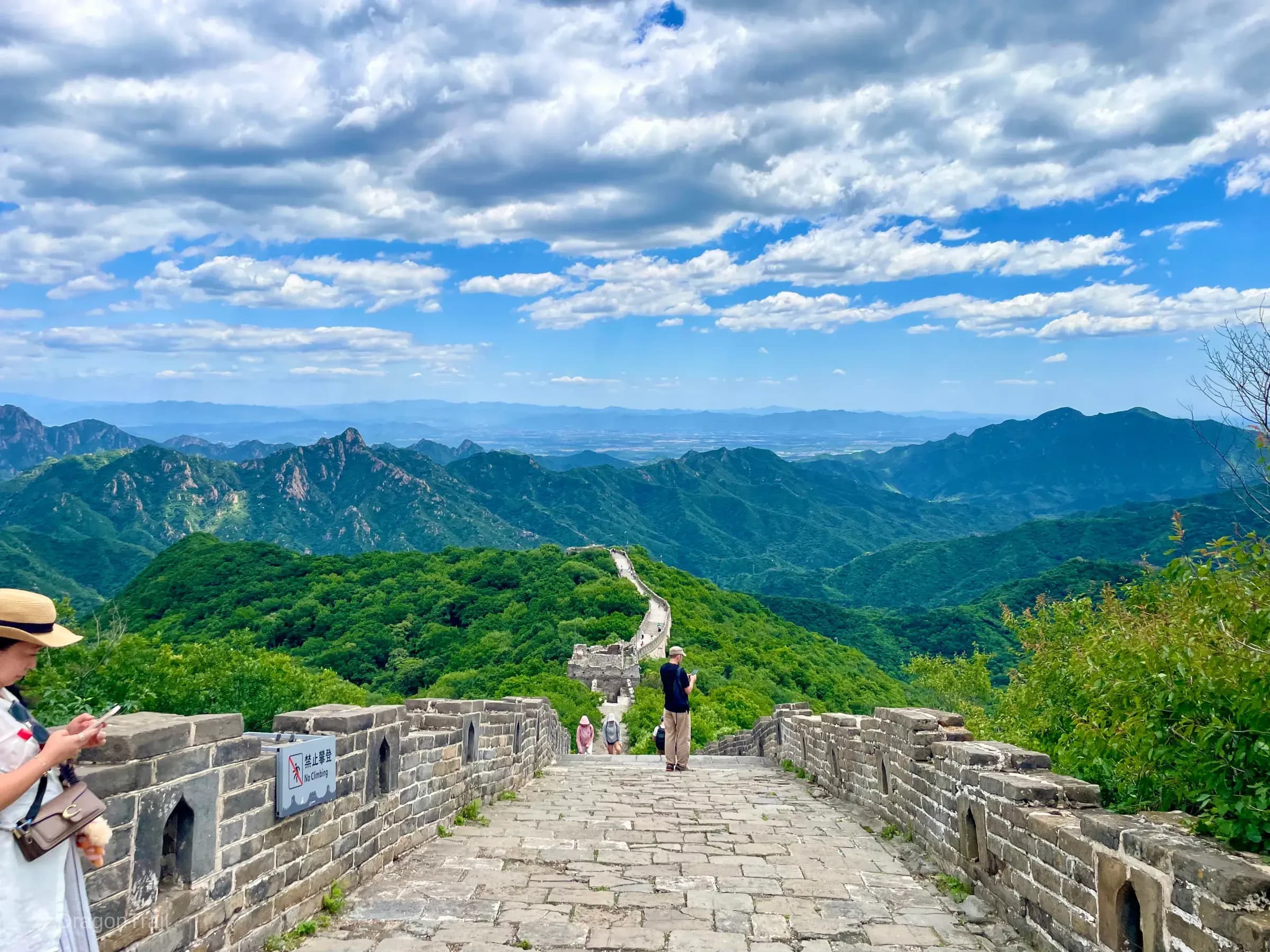Looking down Mutianyu’s steep stone wall path toward a distant watchtower, with crenellated parapets, a no-climbing sign on the left, a visitor in a white dress, and forested mountains under a blue sky