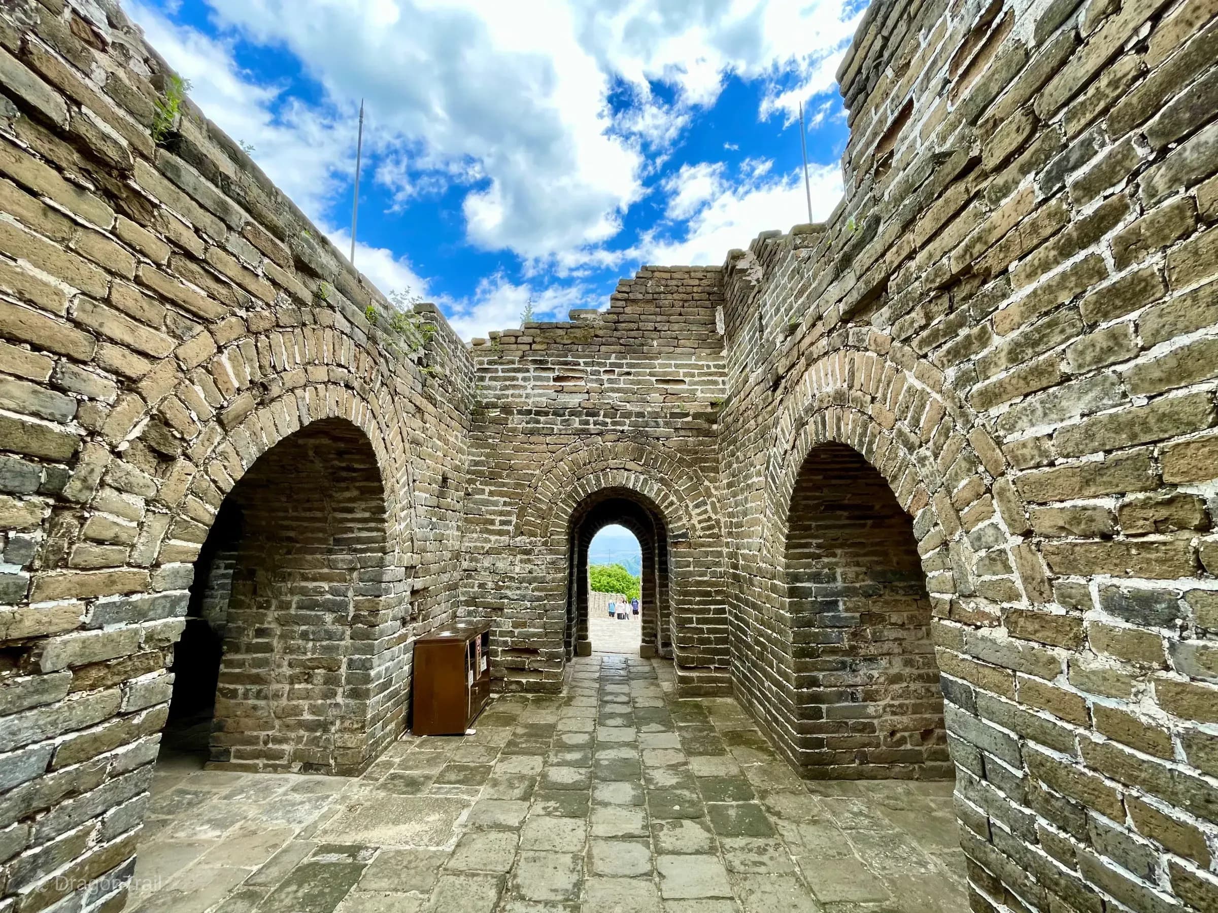 Interior of Mutianyu Tower 22 looking down a stone-paved corridor through three weathered brick arches open to a blue sky with white clouds, with green hills framed by the far doorway