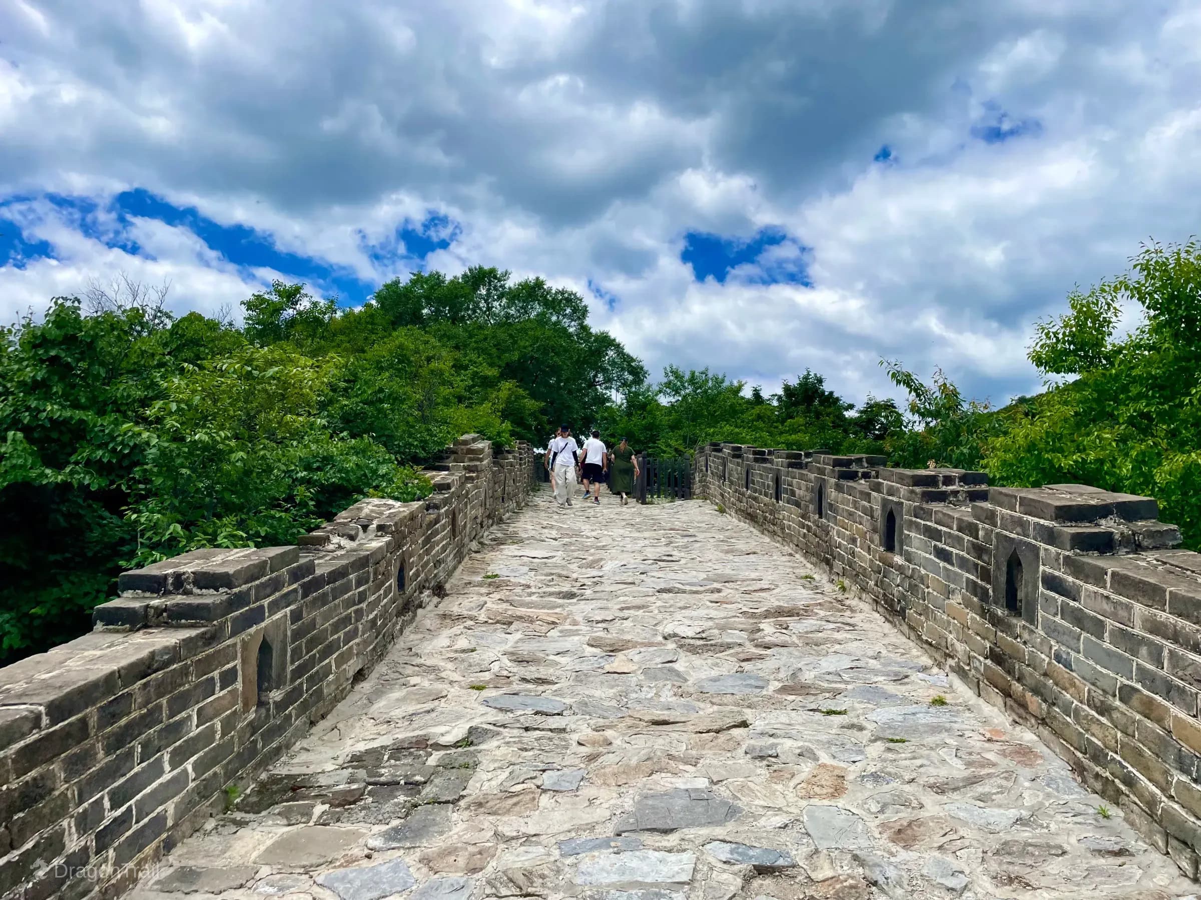 Wide Mutianyu stone wall walk flanked by crenellated parapets and dense green forest, with tourists walking toward a dark metal gate under a dramatic cloudy sky