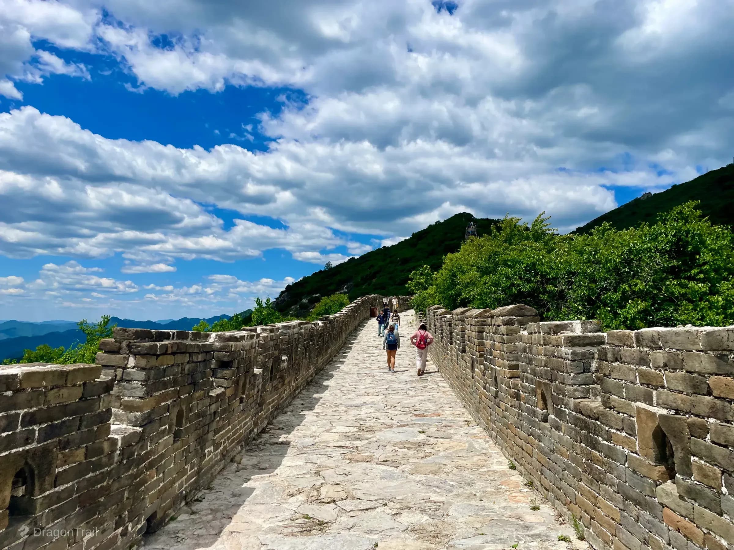 Looking along Mutianyu’s stone wall path with tourists ahead, crenellated parapets, a steep green hillside on the right, distant ridge wall, and layered mountains under a cloudy blue sky