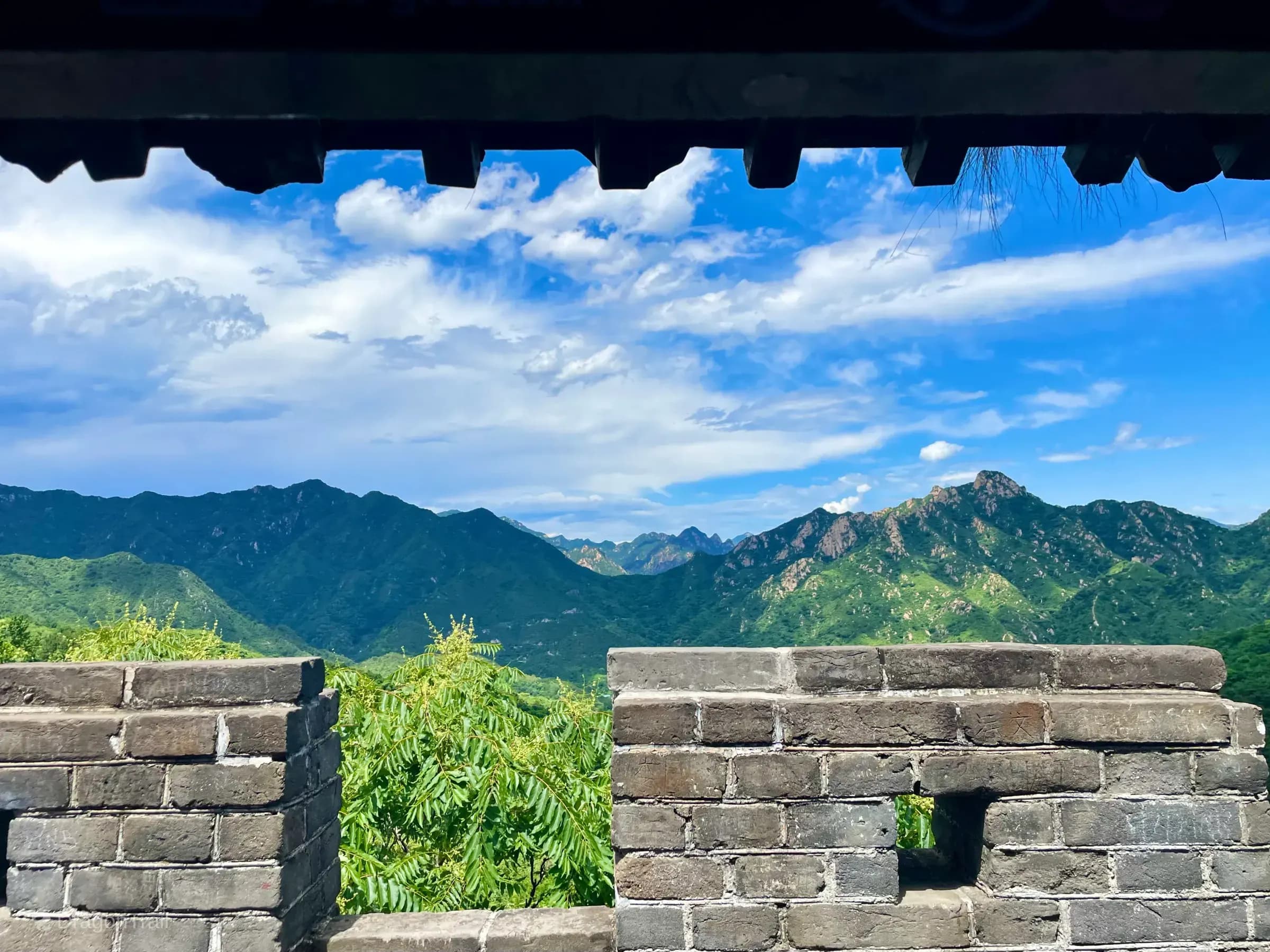 Forested Huairou mountains seen over Mutianyu battlements from beneath a watchtower roof with an arrow slit