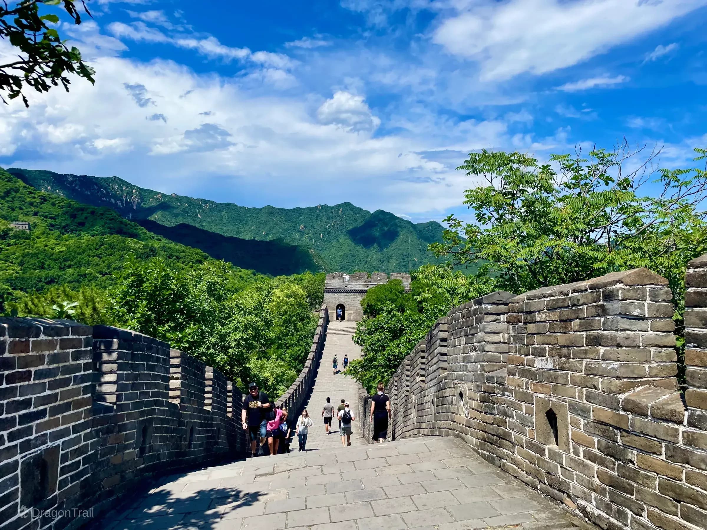 Steep Mutianyu stone walkway descending between crenellated brick parapets toward a rectangular watchtower amid forested Huairou mountains