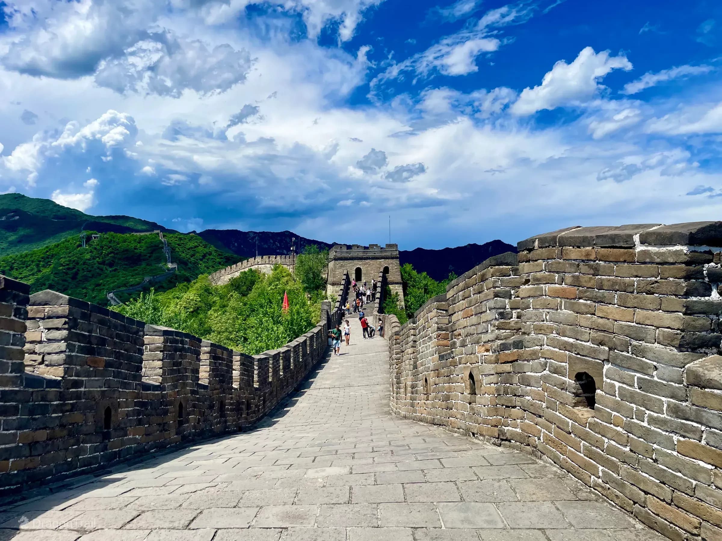 Looking up the Mutianyu wall toward Tower 8 with a bright red flag on the left parapet and the ridge wall continuing into forested mountains