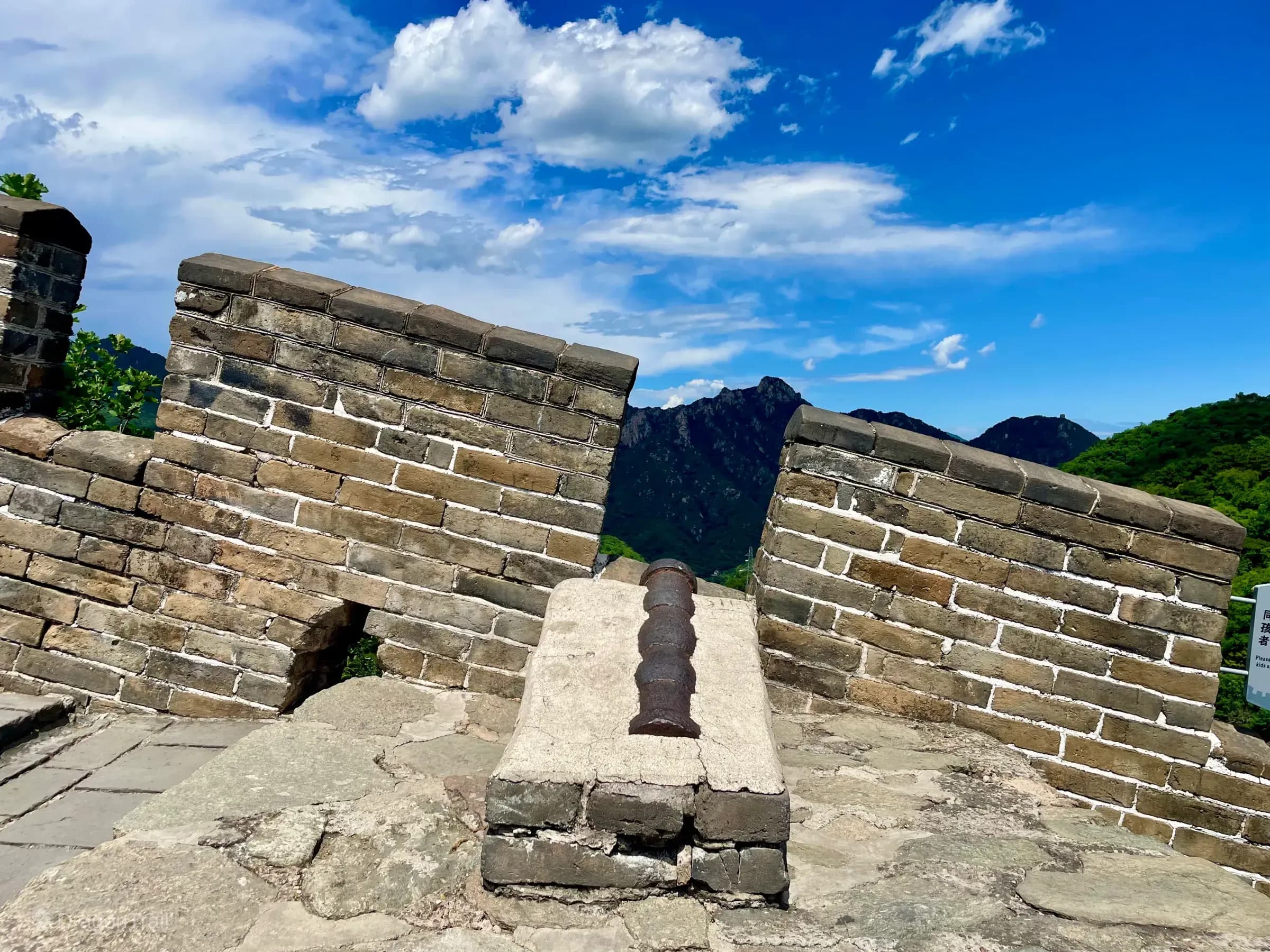 Weathered iron general cannon on a stone pedestal aimed through Mutianyu battlements toward densely forested mountains