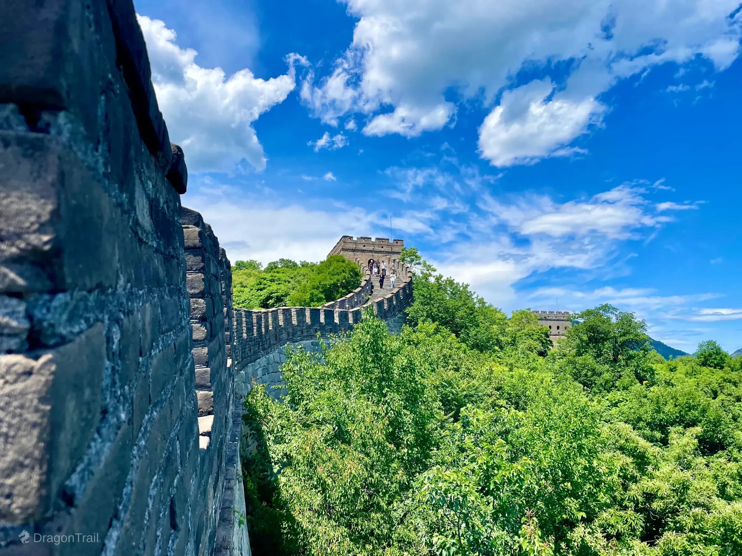 Mutianyu Great Wall climbing a forested ridge toward Tower 9 on a high point, with battlements in the left foreground under dramatic clouds