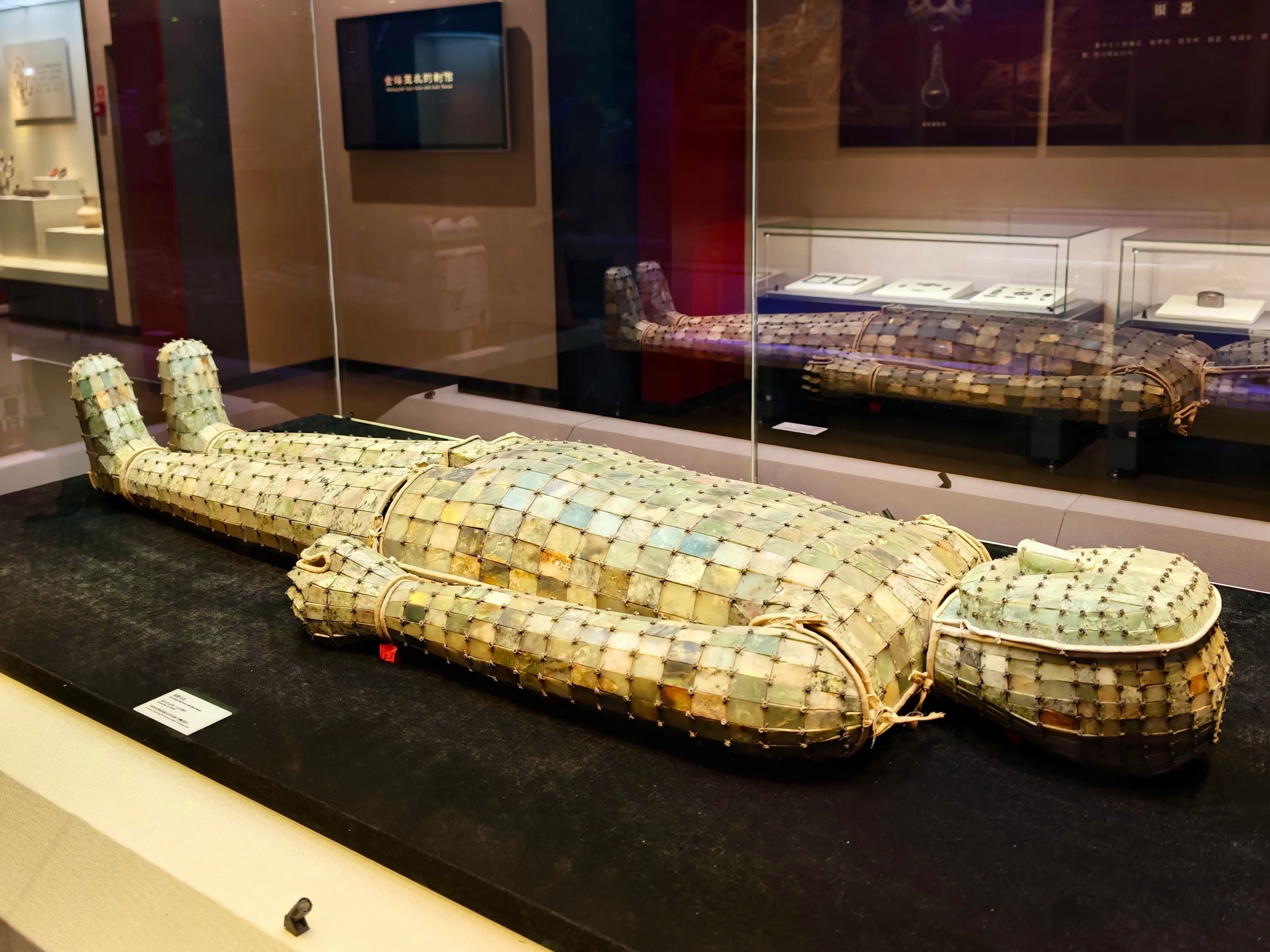 A jade burial suit displayed in a museum case, composed of numerous small jade tiles connected by metal wires.