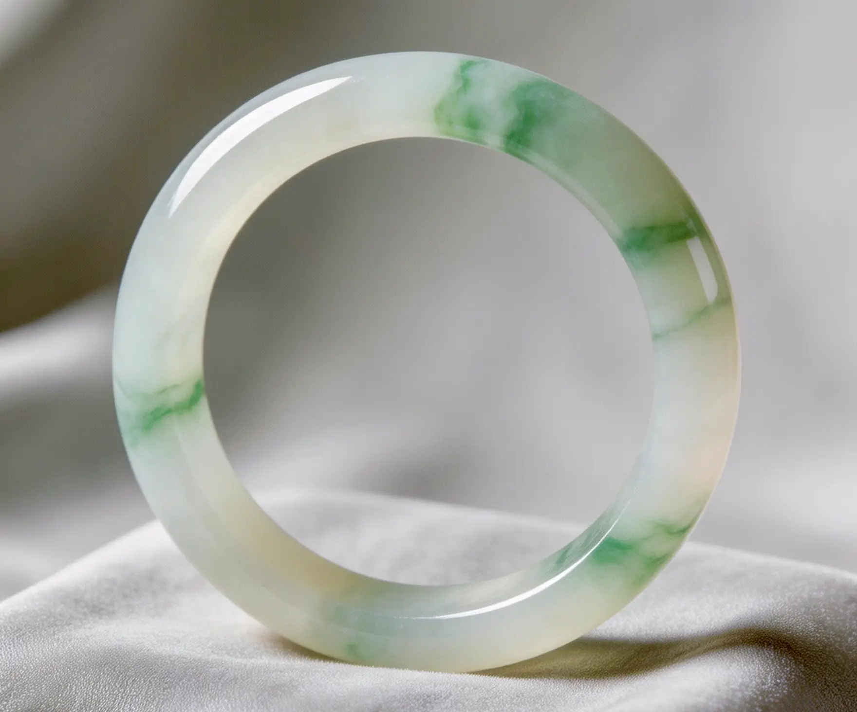 A jade bangle bracelet with green and white hues displayed on a soft fabric.