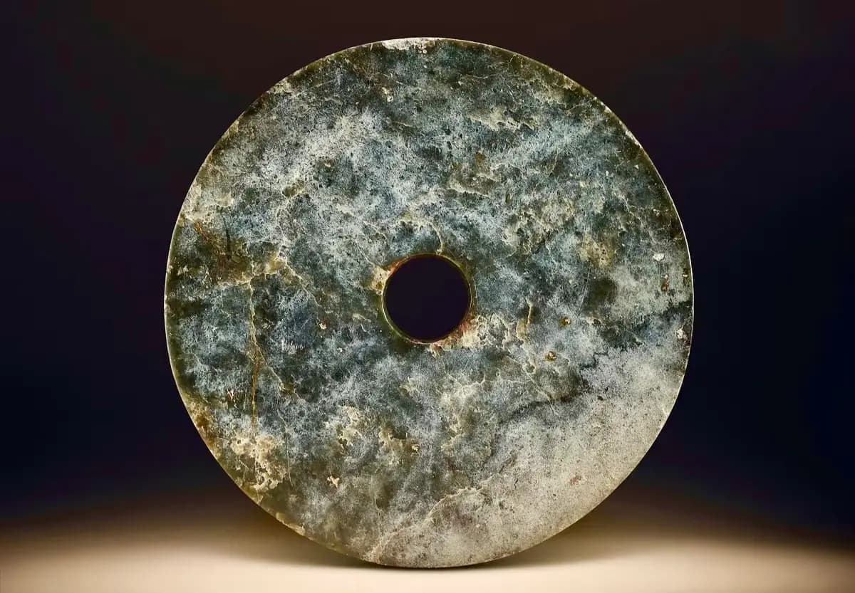 A circular, flat, jade disc with a central hole, exhibiting a mottled grey and green surface.