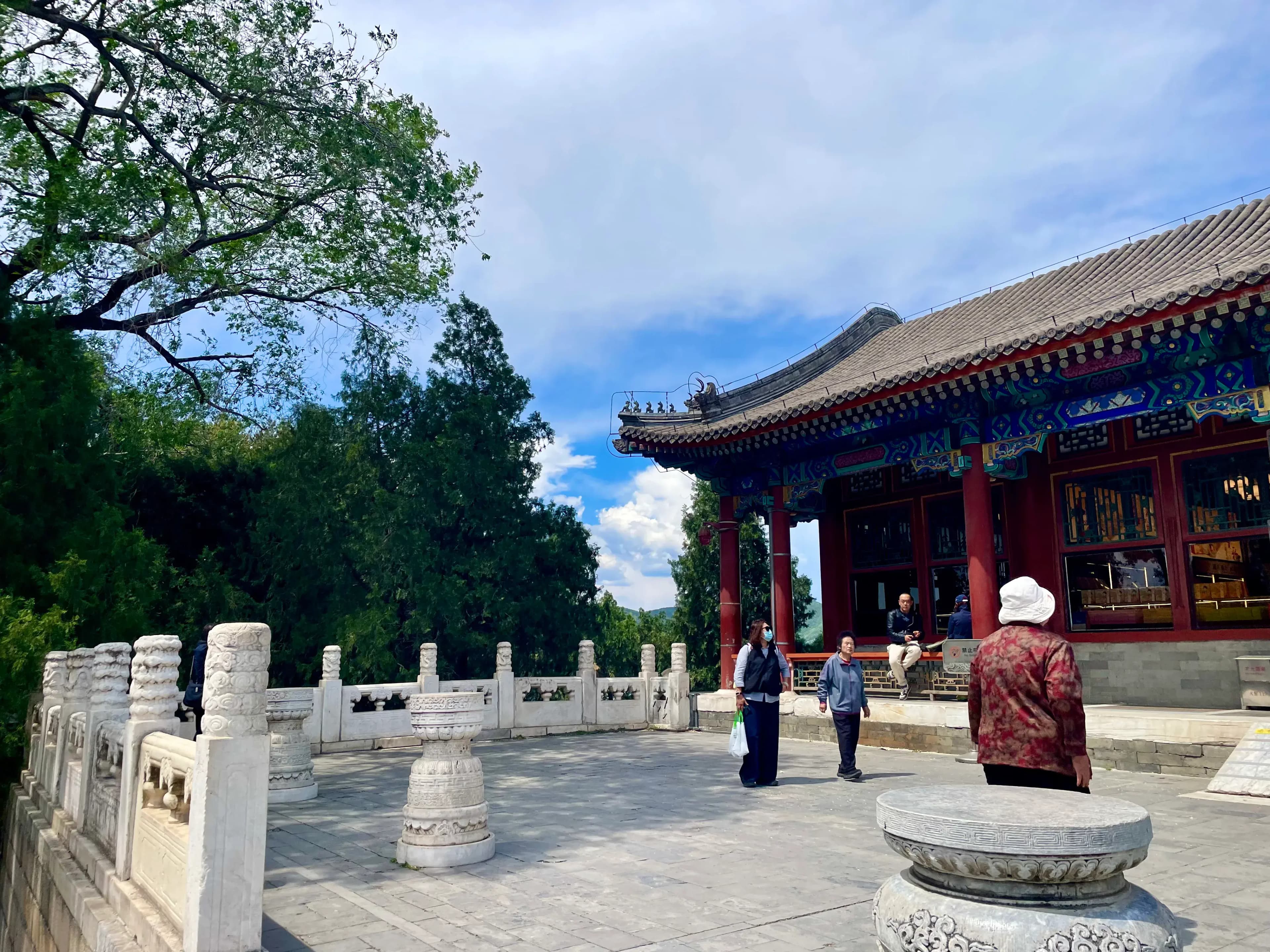 A courtyard with traditional Chinese architecture, featuring a building with ornate eaves and a stone railing. Several people are walking around the area.