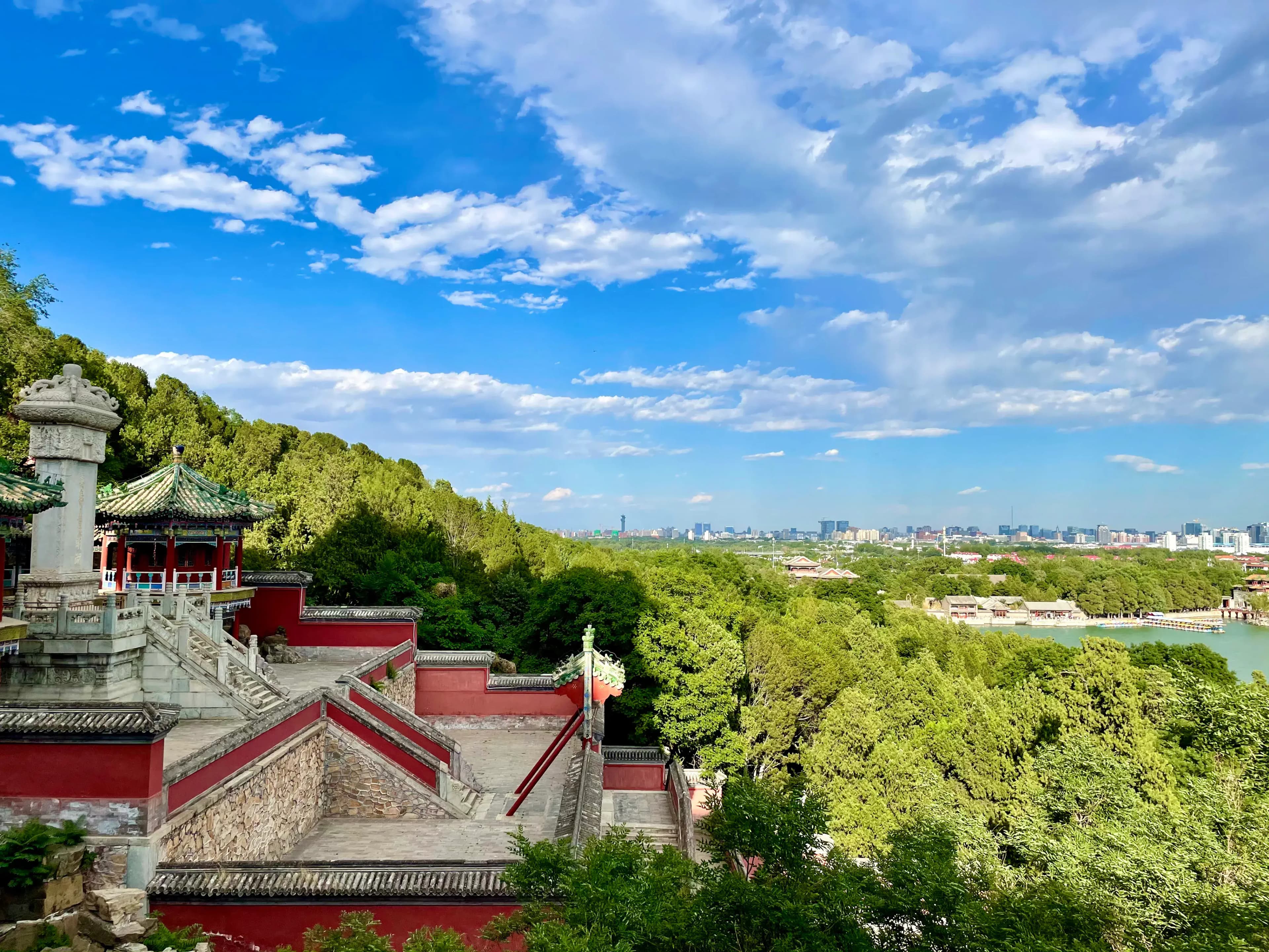 A traditional Chinese pavilion with green tiled roof and red walls, surrounded by lush green trees and overlooking a cityscape in the distance.