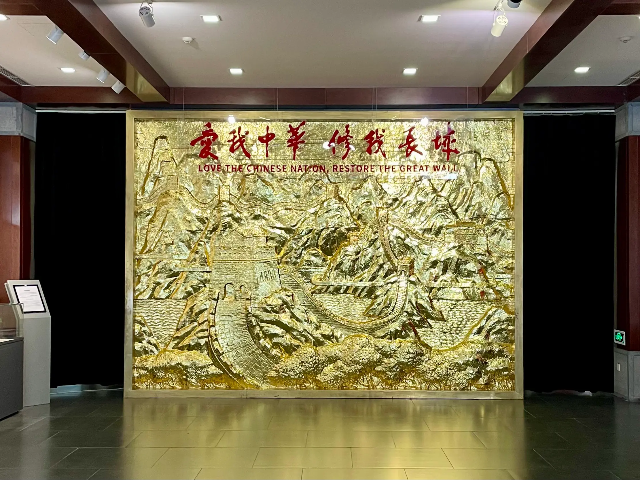 Large gilded relief mural inside Mutianyu's Great Wall Spirit Exhibition Hall, depicting the Ming Great Wall winding across mountain ridges beneath the bilingual campaign slogan “Love the Chinese Nation, Restore the Great Wall.”