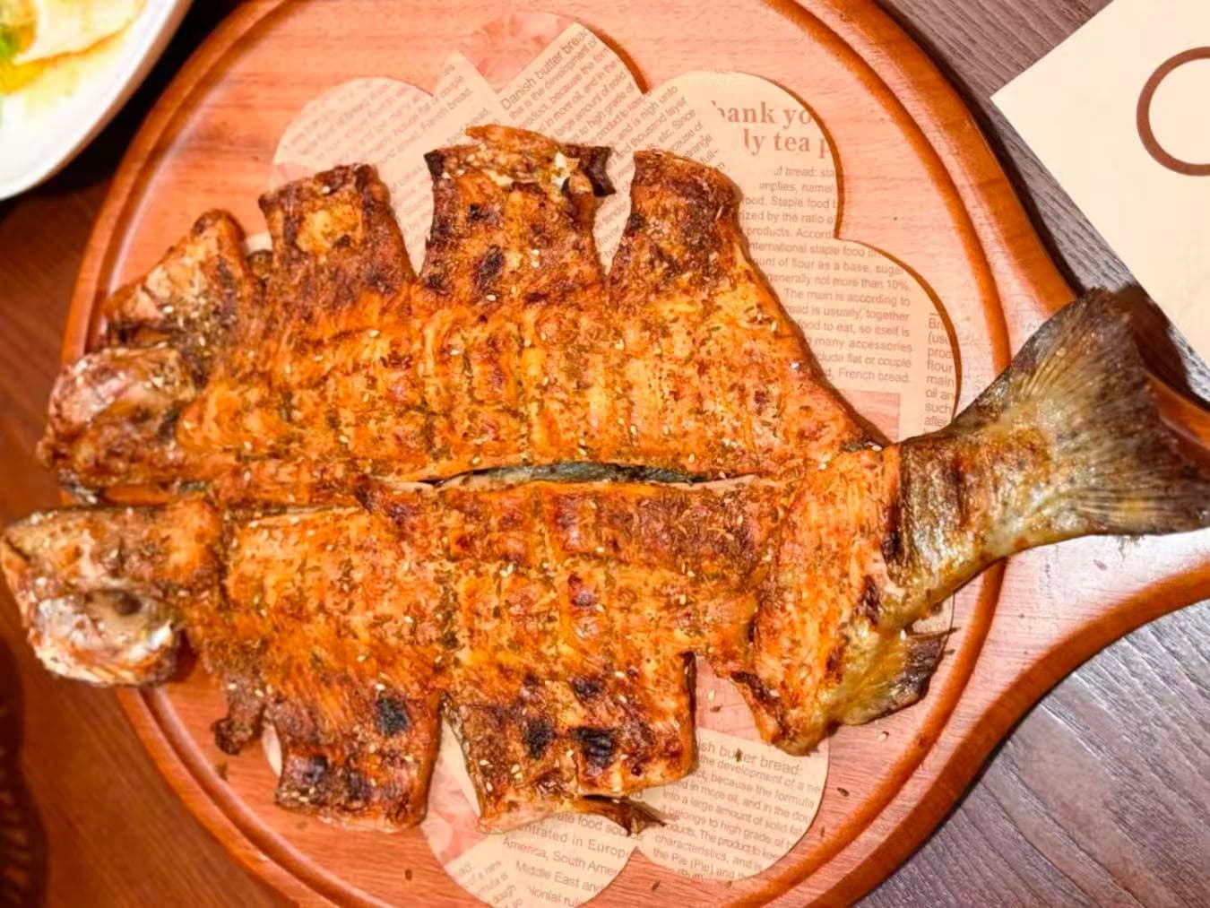 Plated whole Huairou rainbow trout served at a Mutianyu Great Wall scenic-area sit-down restaurant: glossy reddish-brown skin with diagonal scoring marks, head and tail intact, ginger slices on top, presented on a round tan plate on a stone surface — a regional Huairou cold-water fish often served as a signature Mutianyu lunch dish.