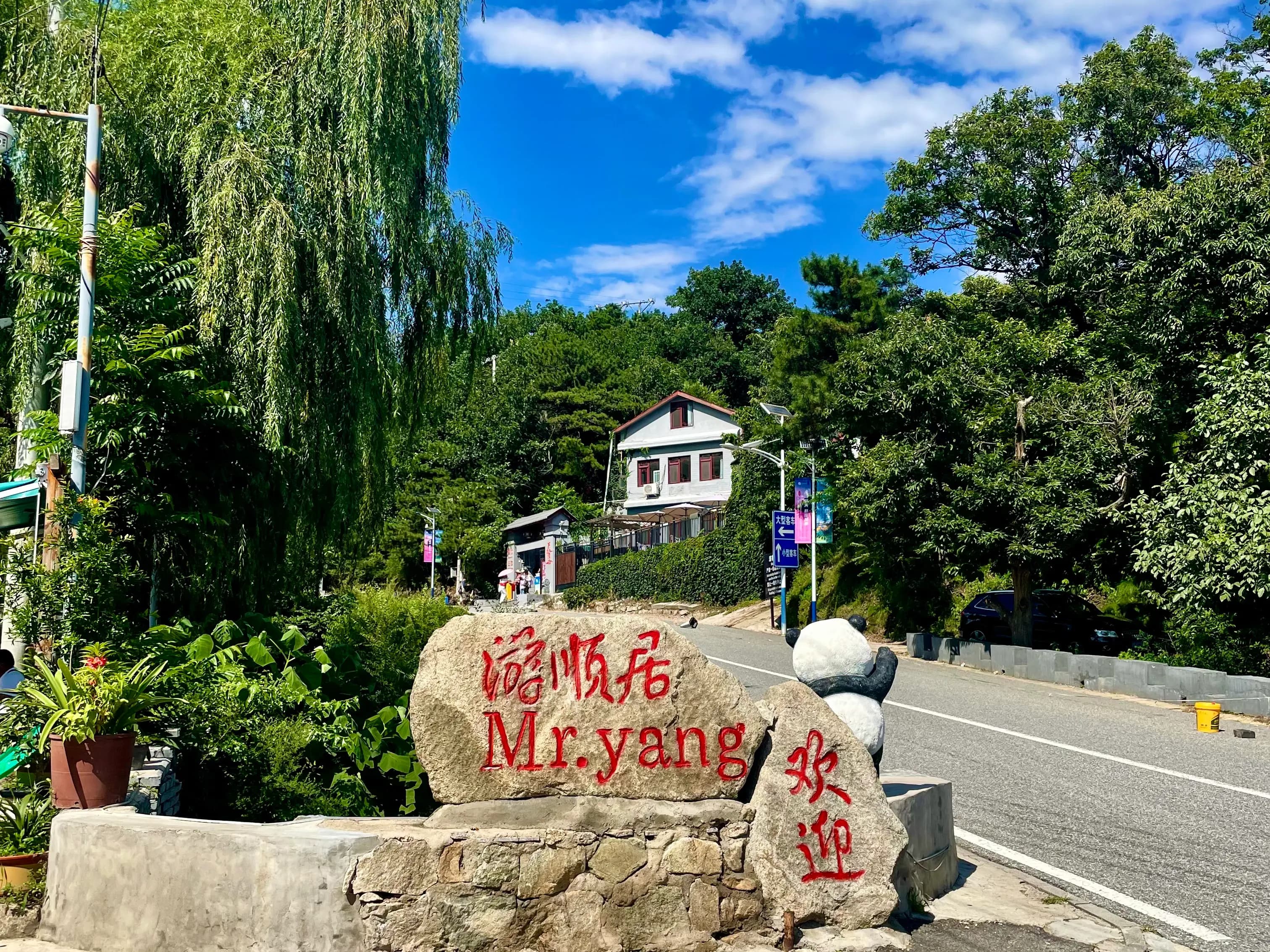 Stone welcome marker on the tree-lined road into Mutianyu village, with a hilltop guesthouse visible behind willow trees under a blue sky.