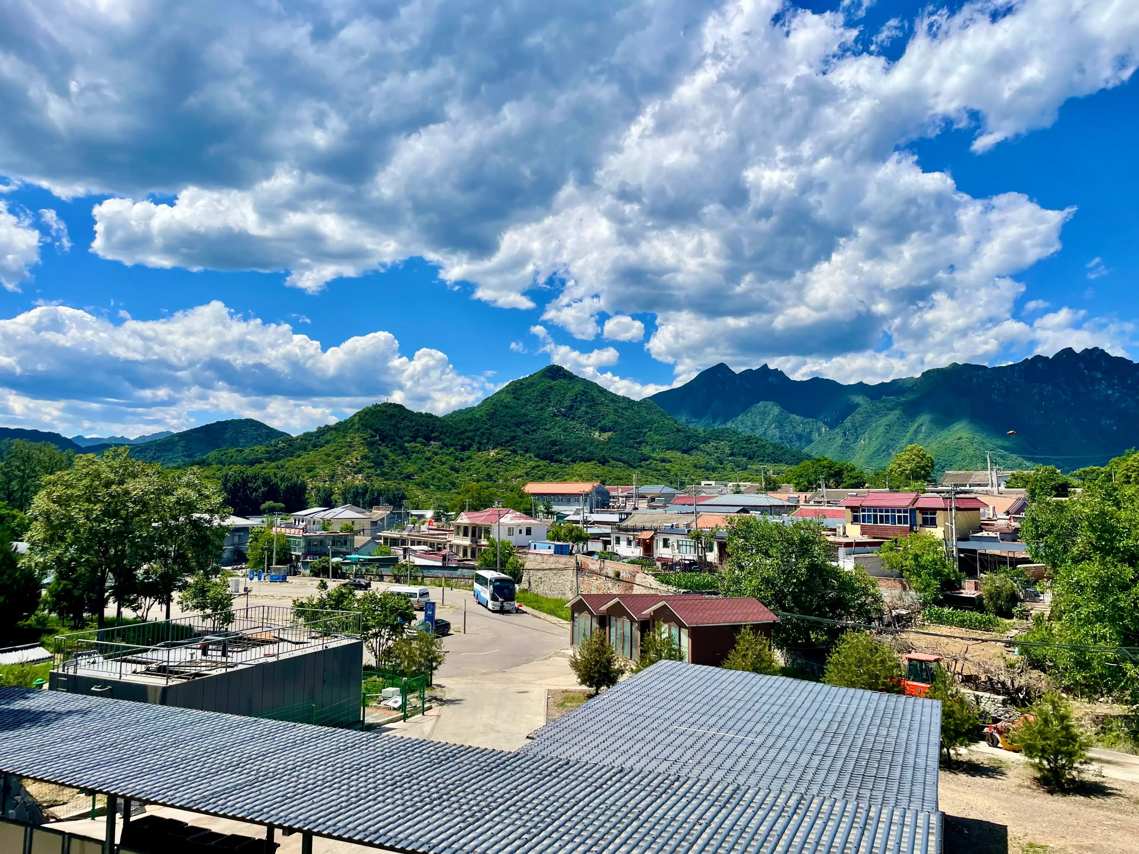 Rooftops of Xinying village near Mutianyu, with the surrounding mountain ridgeline rising in the background under a blue sky, showing Xinying’s position east of Mutianyu and below Jiankou as a quieter overnight base.