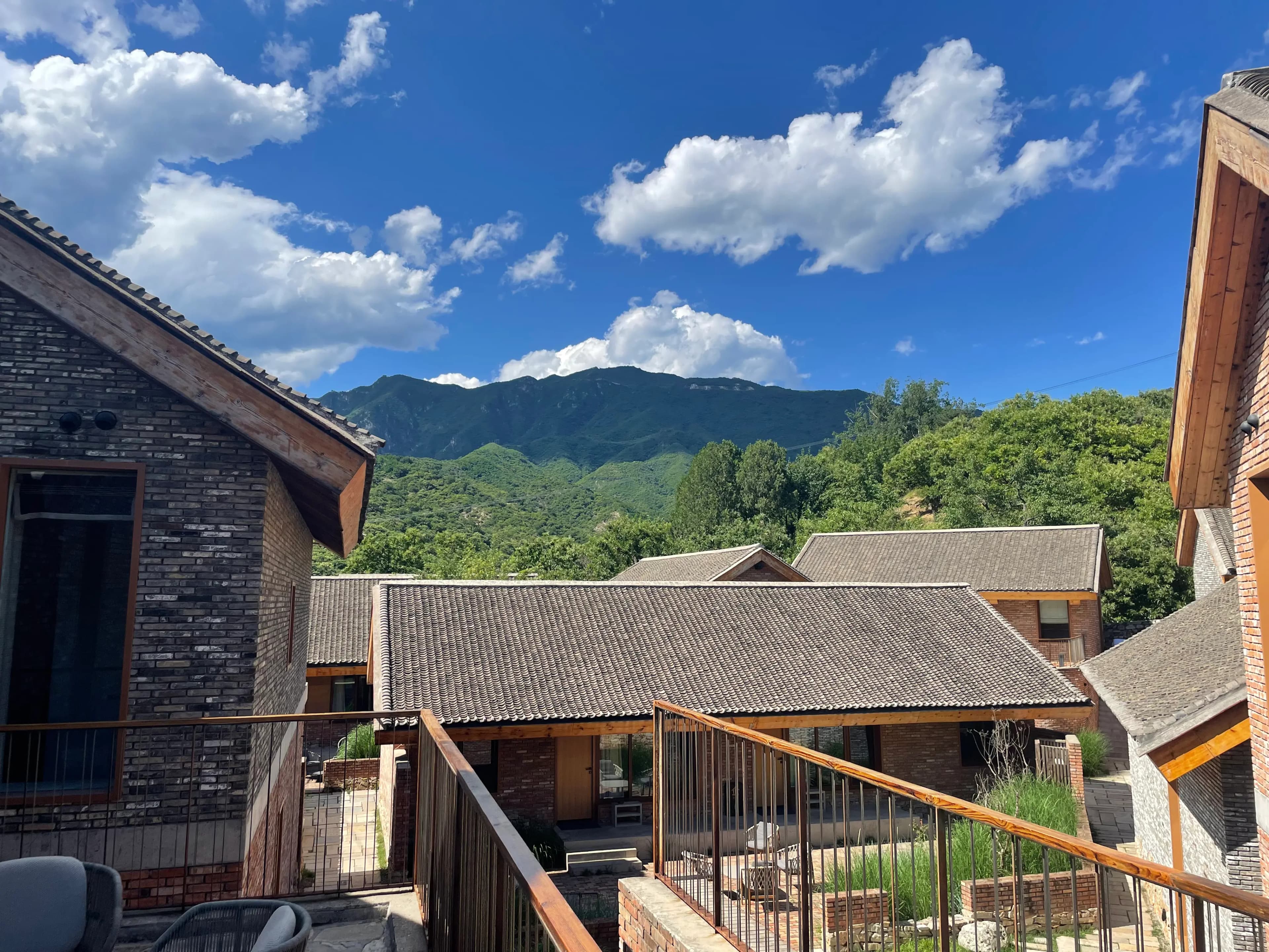 View from a Beigou village courtyard stay near Mutianyu: tile-roofed brick-and-timber houses in the foreground, the upper Mutianyu Great Wall along the ridge near watchtowers 21–23 in the distance, and the historic Cultural Revolution–era hillside slogan “Be loyal to Chairman Mao” still visible on the mountainside.