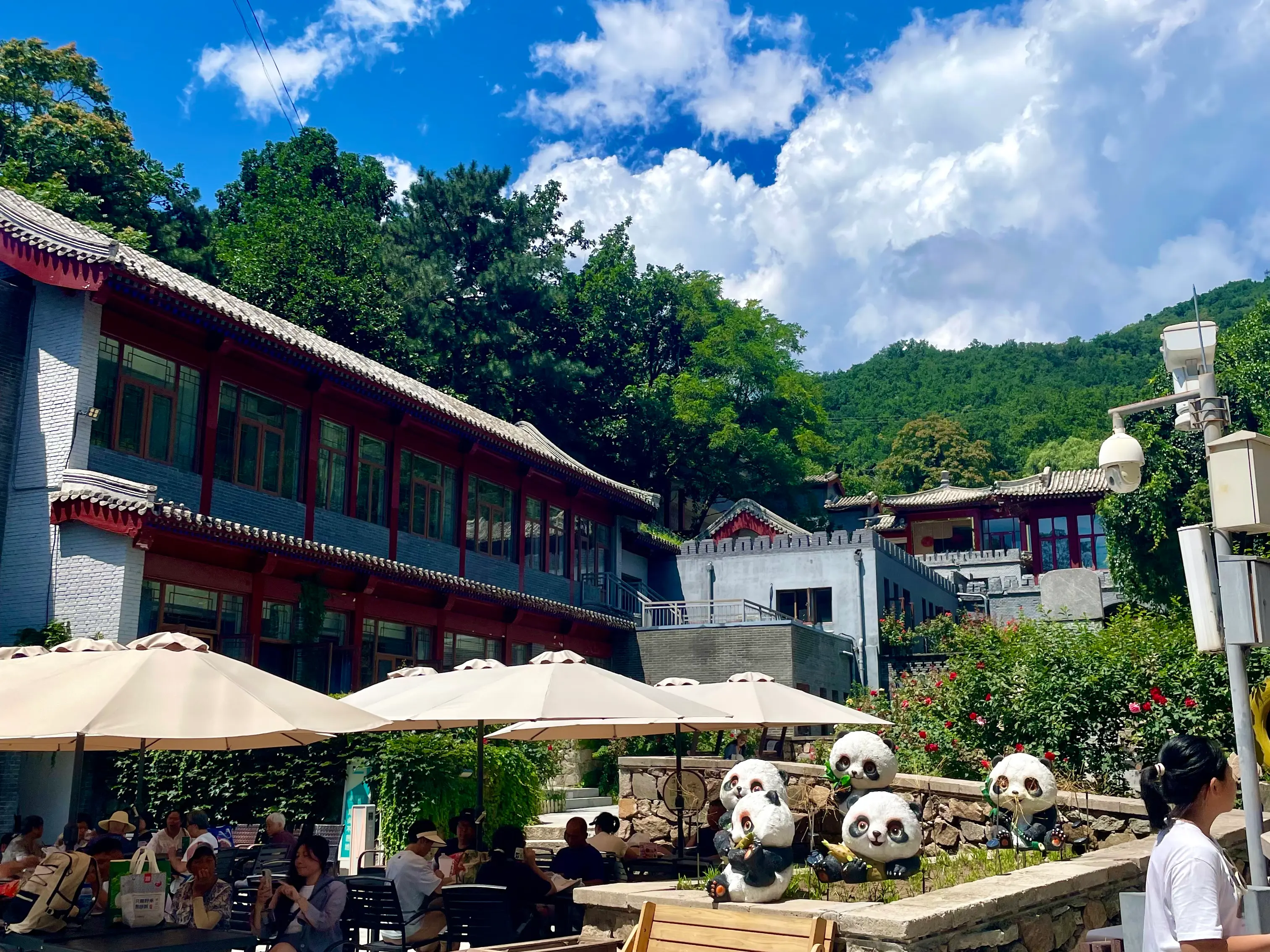 Outdoor café terrace inside the Mutianyu Great Wall scenic area: a two-storey brick-and-tile café building with multiple tan parasols shading café tables on a stone-paved plaza, surrounded by greenery with the Yanshan mountain ridge in the background under a blue sky — a sit-down coffee and drinks option between the visitor gate and the cable-car checkpoint.
