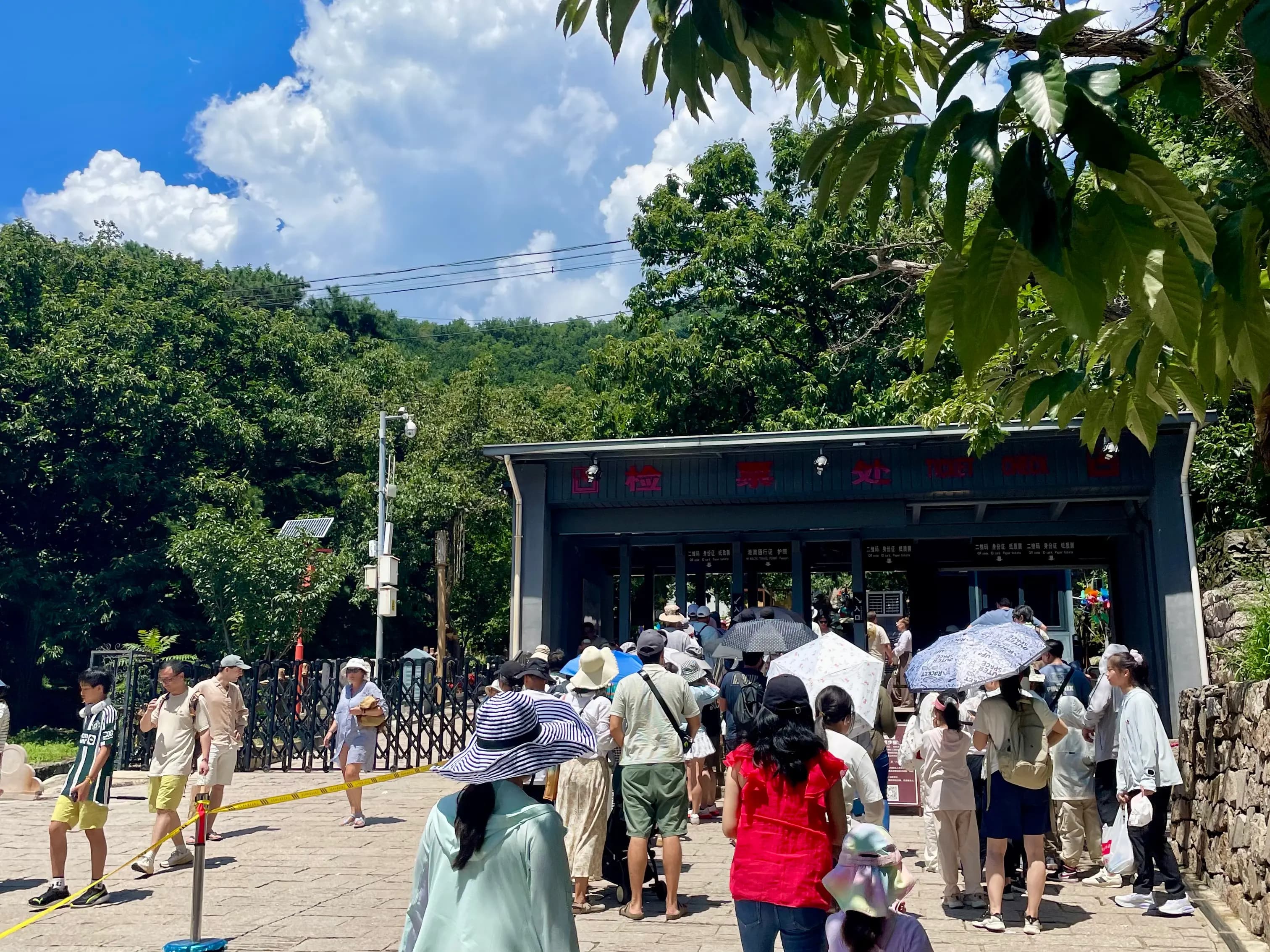 Visitors queuing at the Mutianyu Great Wall west route ticket checkpoint, under a covered structure surrounded by green forest.
