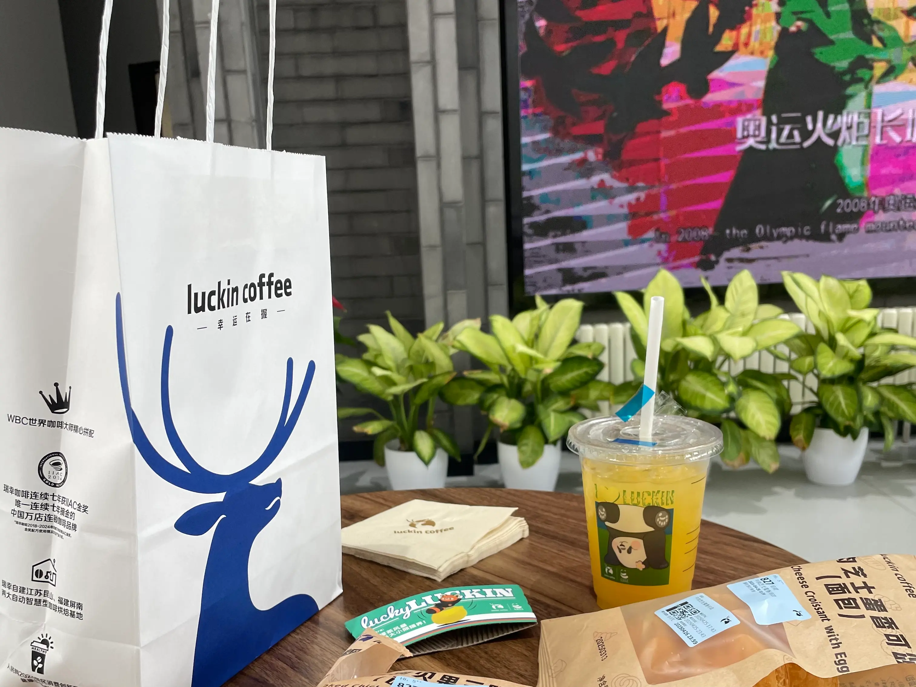 Inside a coffee shop near the Mutianyu Great Wall visitor area: a Luckin Coffee takeaway paper bag with the brand’s blue stag logo, a yellow paper cup, small potted greenery, and a colourful fabric backdrop on a wooden counter — illustrating that mainstream Chinese coffee chains operate takeaway counters inside the scenic-area service zone.