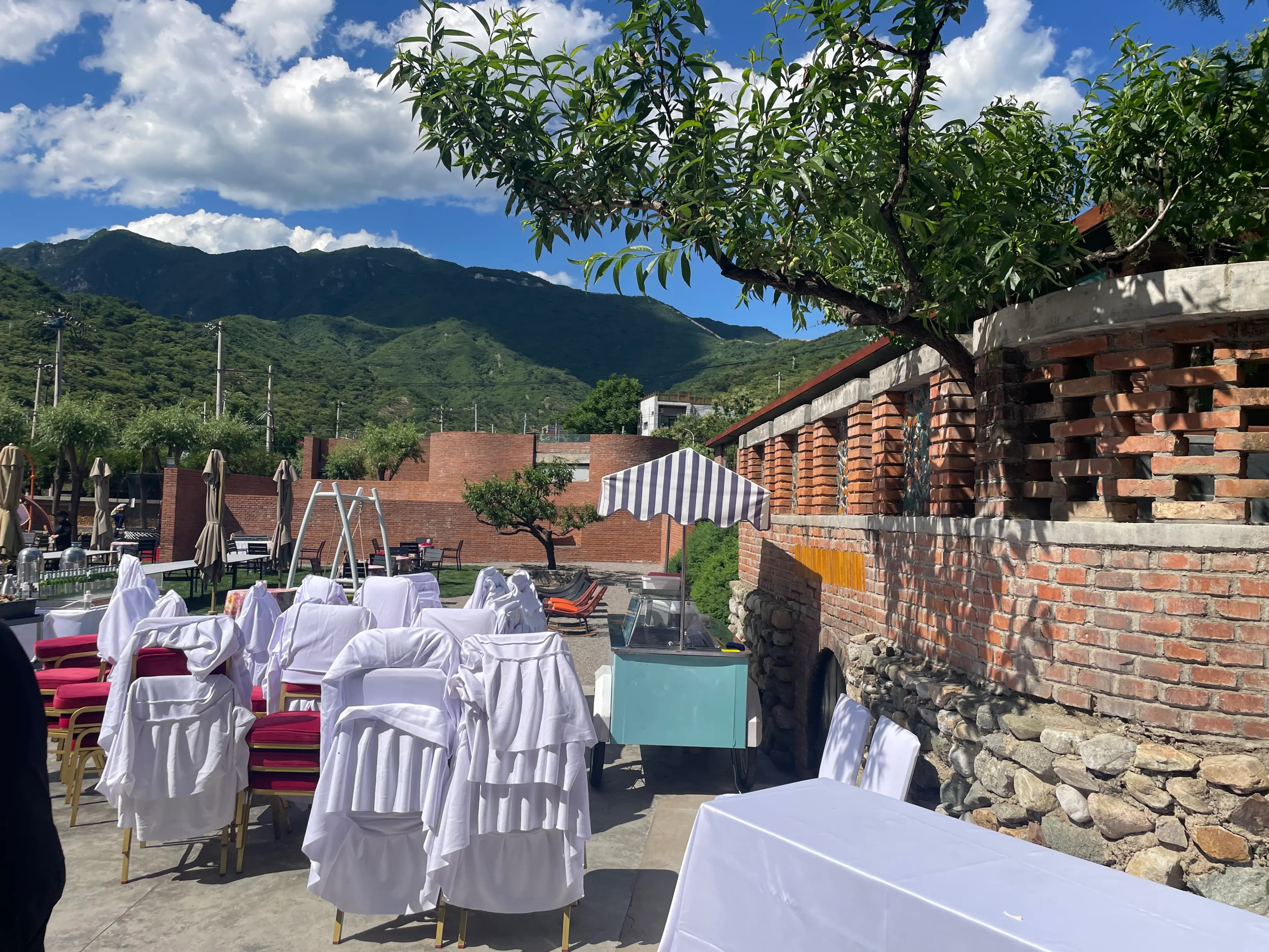 Outdoor terrace at a Beigou-area courtyard restaurant near Mutianyu Great Wall, set up for a banquet: rows of banquet chairs in white cloth covers with red cushions, white tablecloths, a teal mobile buffet cart under a striped parasol, brick walls with decorative inset roof-tile patterns, and the green Yanshan mountain ridge rising under a blue sky.