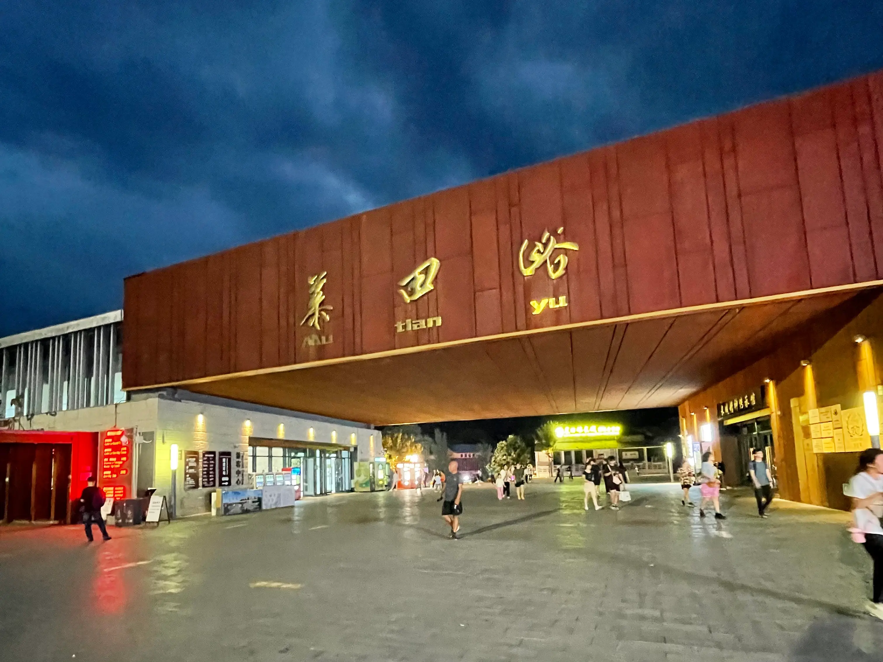Night view of the Mutianyu Great Wall main entrance gateway, with corten-steel cladding lit warmly under a dark blue evening sky and the gold characters Mu tian yu glowing above visitors walking in across wet pavement.