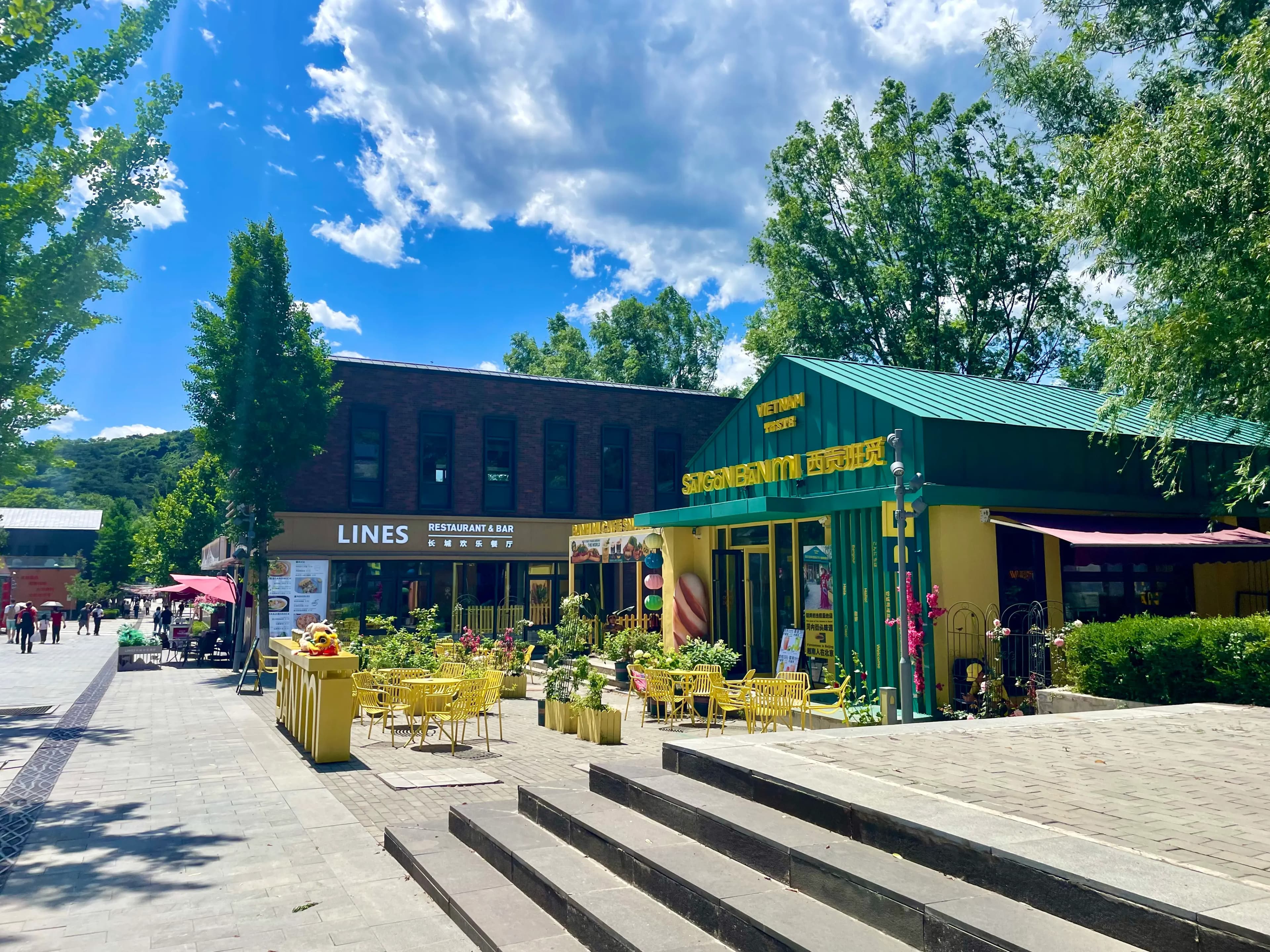 Outdoor dining plaza inside the Mutianyu Great Wall scenic-area arcade between the main entrance and the ticket / shuttle / cable-car checkpoints, with a brown-brick "LINES Restaurant & Bar" storefront and an adjoining green-roofed yellow building branded "Saigon Venture / Vietnam Taste," yellow café tables and chairs, yellow planters with flowers, broad stone steps, and tall green trees under a blue sky with summer clouds.