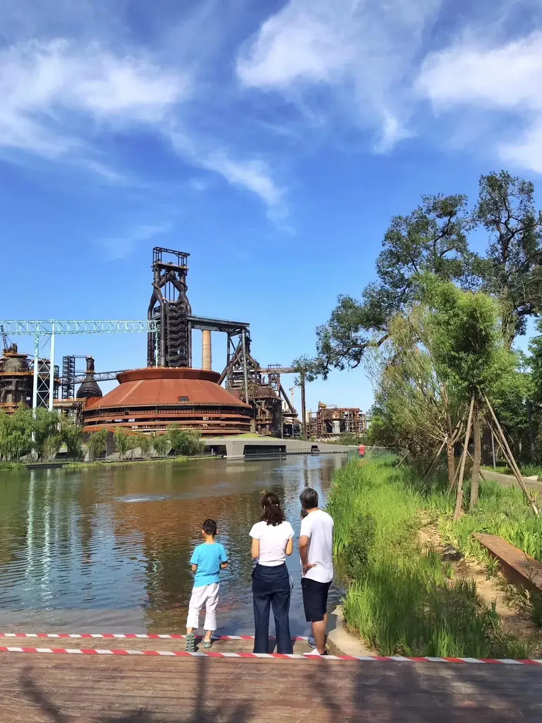 A family stands on a wooden deck looking across the water toward the blast furnace at Shougang Park, Beijing